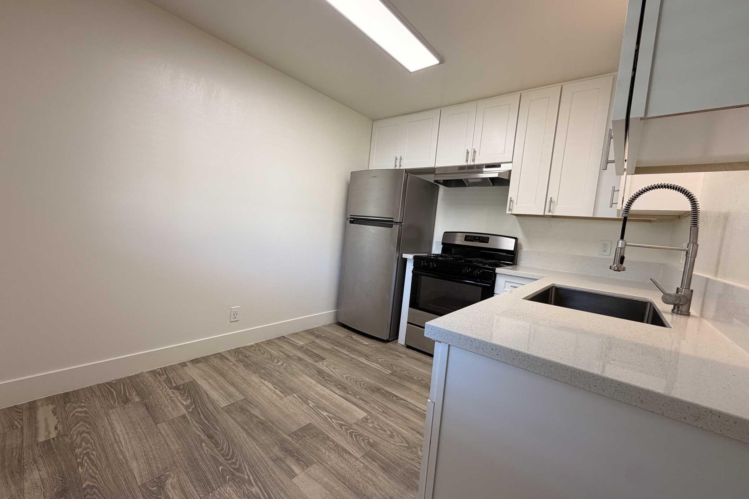 A modern kitchen featuring stainless steel appliances, including a refrigerator and a gas stove, white cabinetry, and a spacious countertop. The room has light-colored walls and a wood-like floor, providing a clean and bright atmosphere. A window provides natural light.