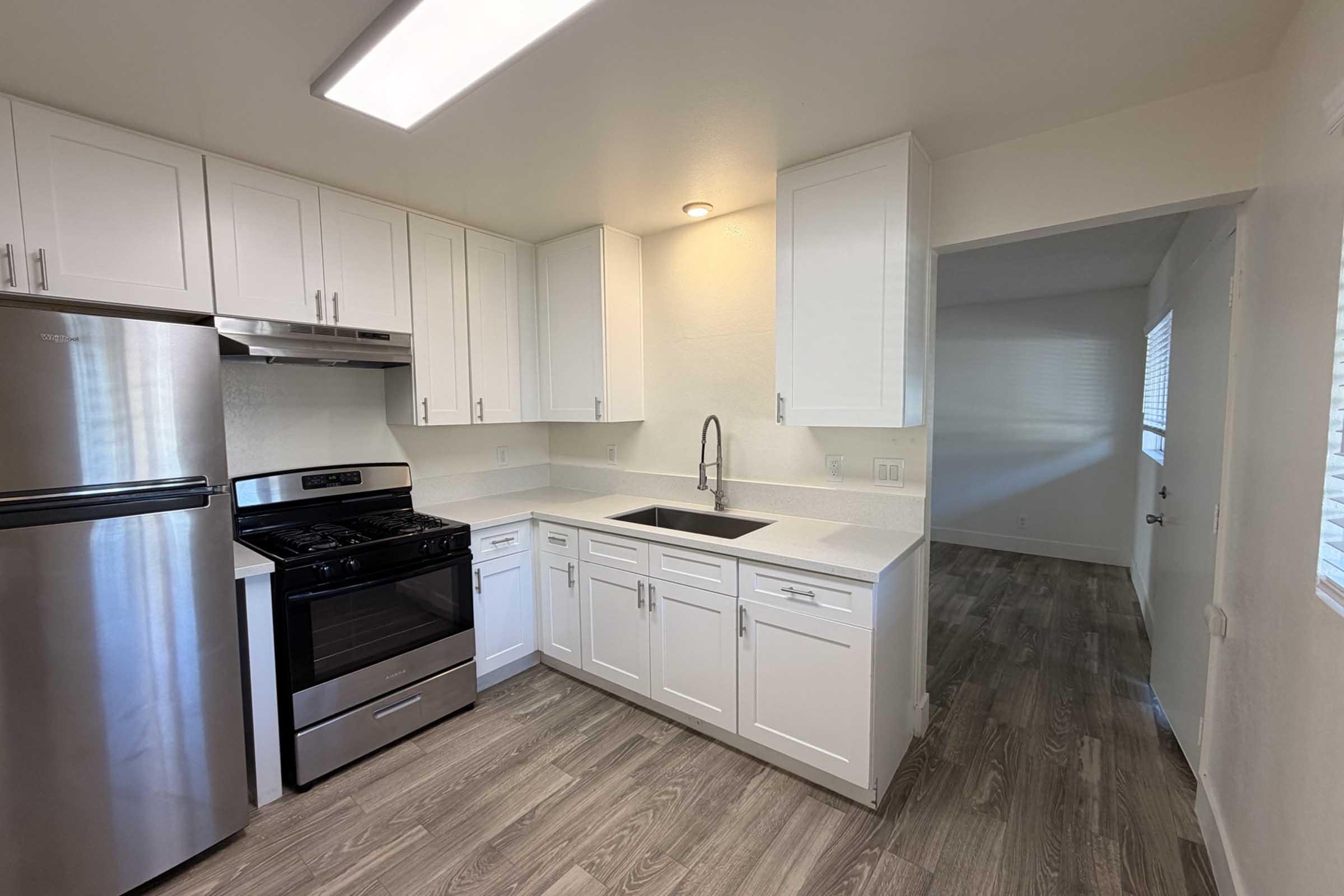 Modern kitchen with stainless steel appliances, including a gas stove and refrigerator. Features white cabinetry, a single basin sink with a sleek faucet, and light-colored countertops. The flooring is a warm wood look, and there’s a doorway leading to a bright, connected living space with window light.