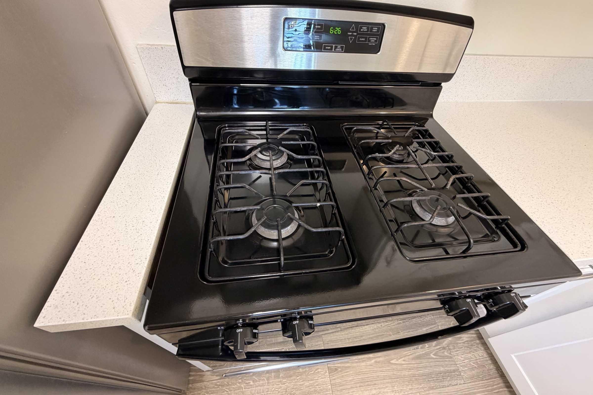 A modern black gas stove with four burners and stainless steel accents, set on a light-colored countertop. The control panel displays digital settings, and the stove is positioned against a beige wall. The surrounding area is clean and organized, giving a contemporary kitchen vibe.