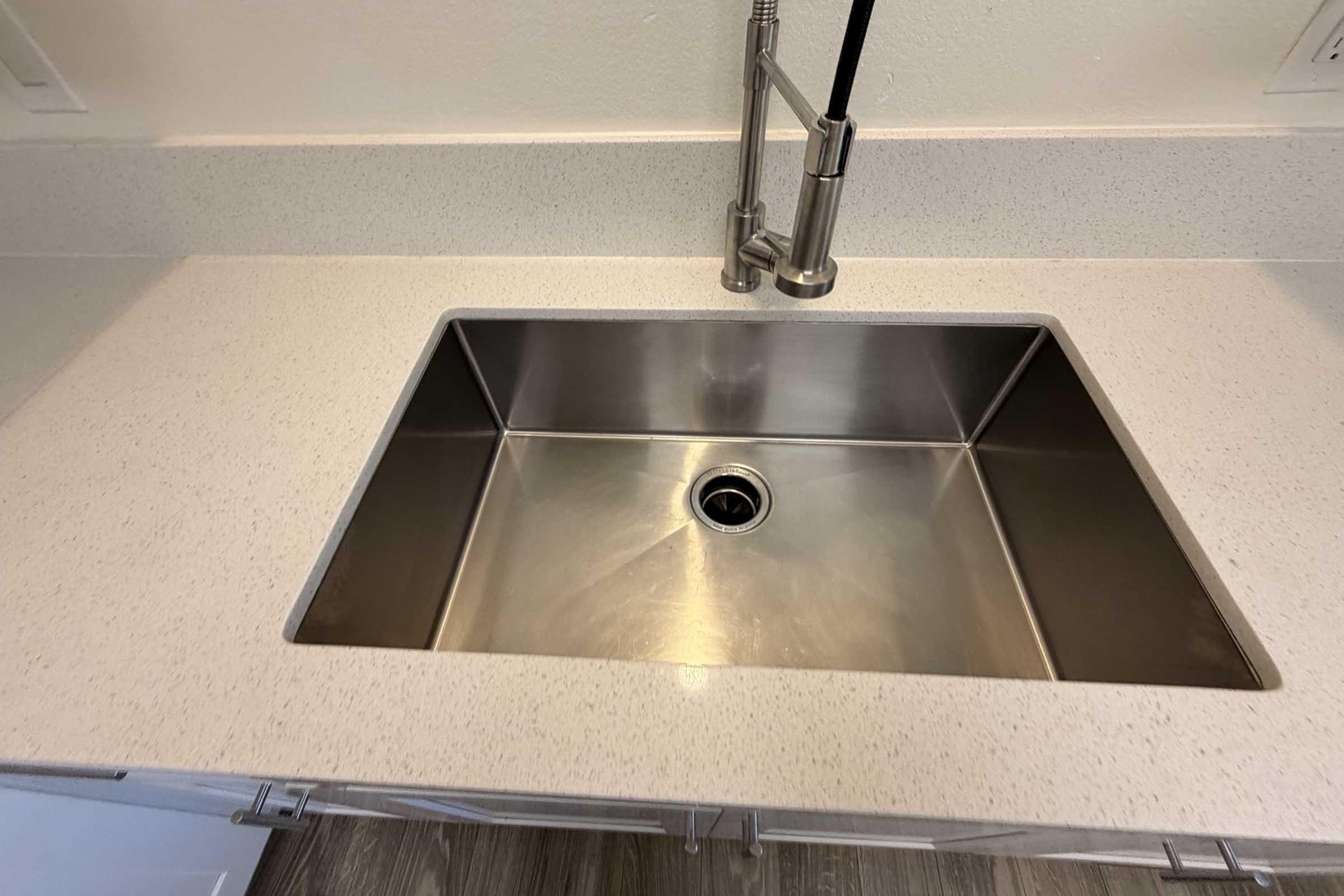 A clean stainless steel kitchen sink set into a light-colored speckled countertop. The sink features a single drain in the center and is surrounded by a pristine, modern kitchen space with minimalistic cabinetry.