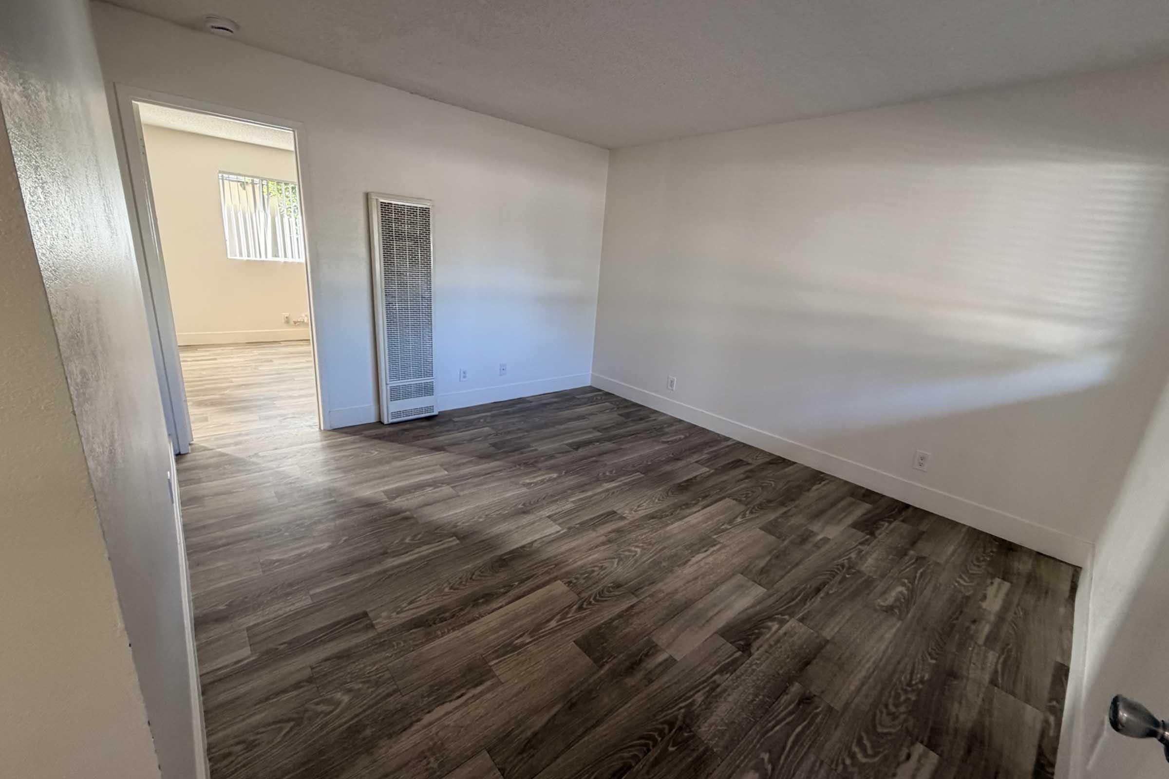 Empty room with light-colored walls and modern wood-like flooring. A heater is mounted on the wall, and a doorway leads to another room. Natural light streams in through a window, creating a bright and open atmosphere. The space is clean and unfurnished, ready for decoration or furnishing.