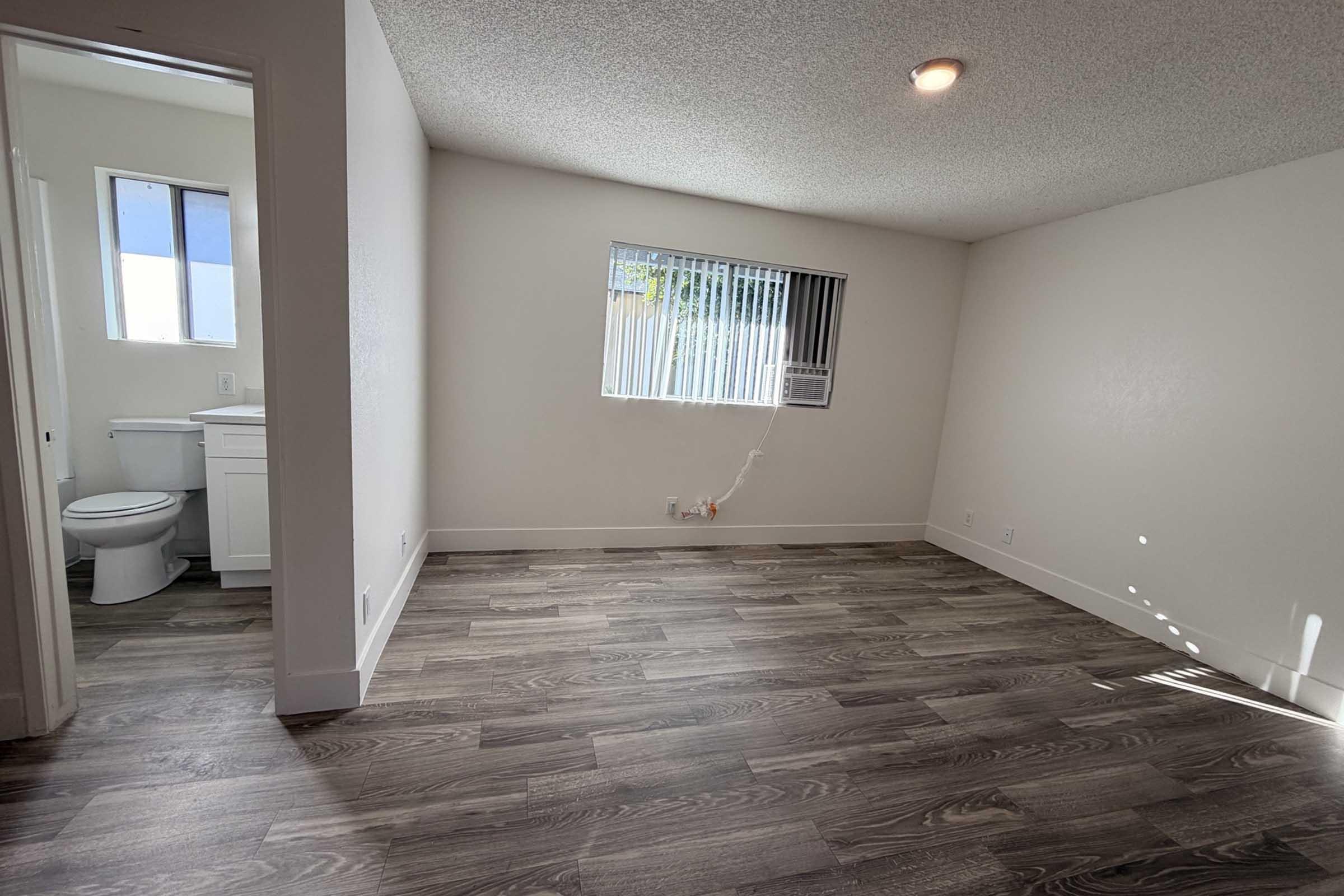 A bright, empty room with wooden laminate flooring and white walls. A small window with vertical blinds lets in natural light. To the left is a bathroom door, and a compact bathroom features a toilet and sink. An air conditioning unit is mounted in the wall beneath the window.