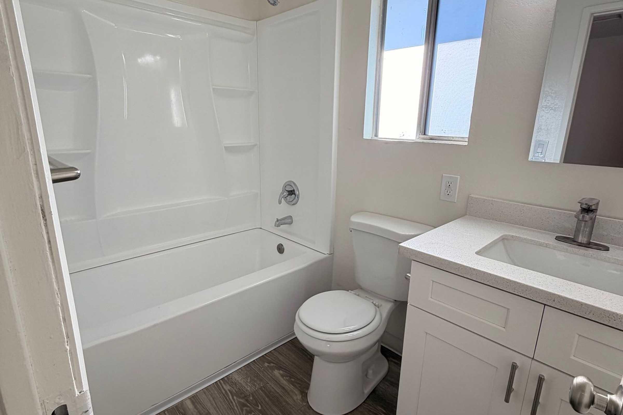 A well-lit bathroom featuring a white bathtub with a shower, a white toilet, and a modern sink with a countertop. The walls are painted light, and there's a window allowing natural light. The flooring is a wood-like laminate, adding a modern touch to the space.