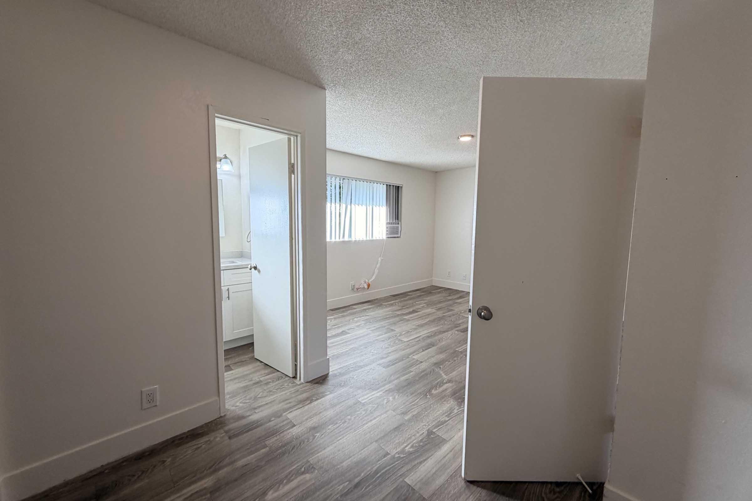 Interior view of a room featuring a doorway leading to a bathroom. The room has light-colored walls, textured ceiling, and wood-like flooring. There is a window with blinds letting in natural light, and an open door reveals additional space. The overall atmosphere is clean and well-lit.