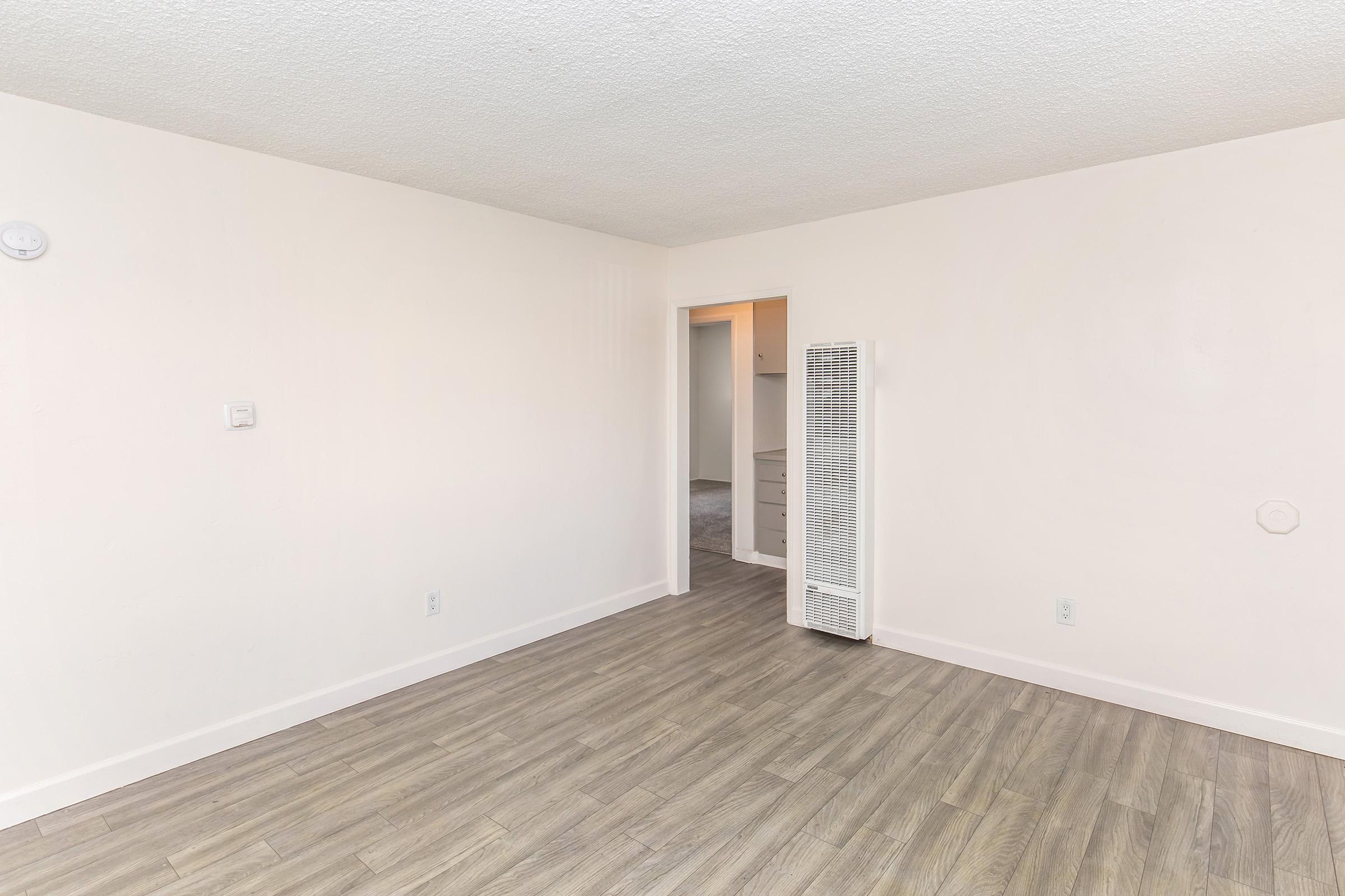 A spacious, empty room with light-colored walls and gray laminate flooring. A wall-mounted heater is visible on one side, and a doorway leads to another area, suggesting additional space beyond. The overall atmosphere is bright and minimalistic, suitable for various interior designs.