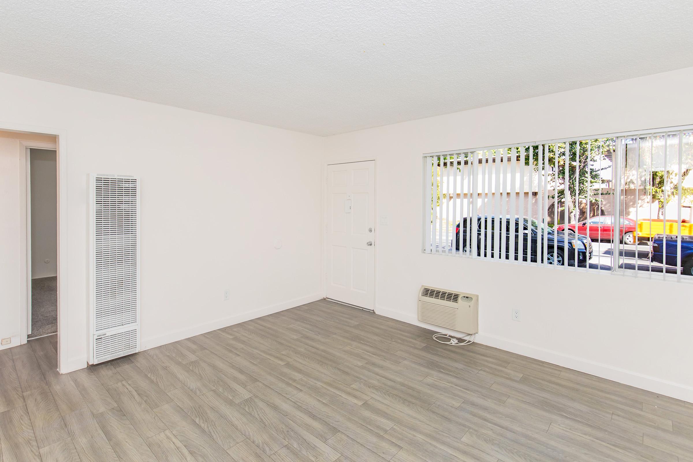 A spacious room with light-colored walls and wooden flooring. There's an air conditioning unit on the wall, a door leading to other areas, and a window with blinds allowing natural light. Outside the window, there are cars parked on the street. The overall aesthetic is clean and minimalistic.