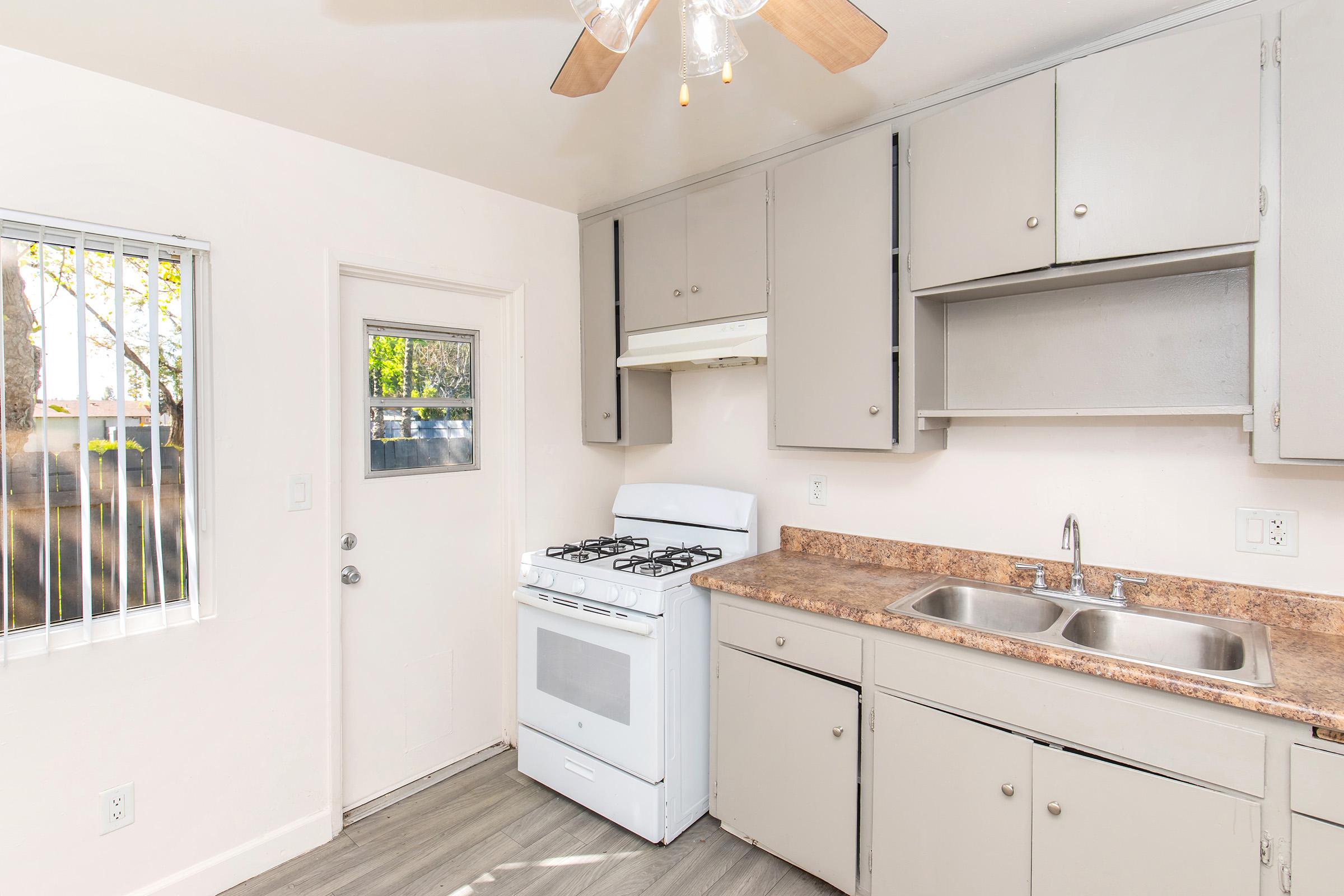 A small, light-colored kitchen featuring a gas stove, sink with two basins, and upper cabinets. There's a window with blinds and a door leading outside. The flooring is a light gray wood-style. The overall look is simple and functional, with neutral colors and adequate natural light.