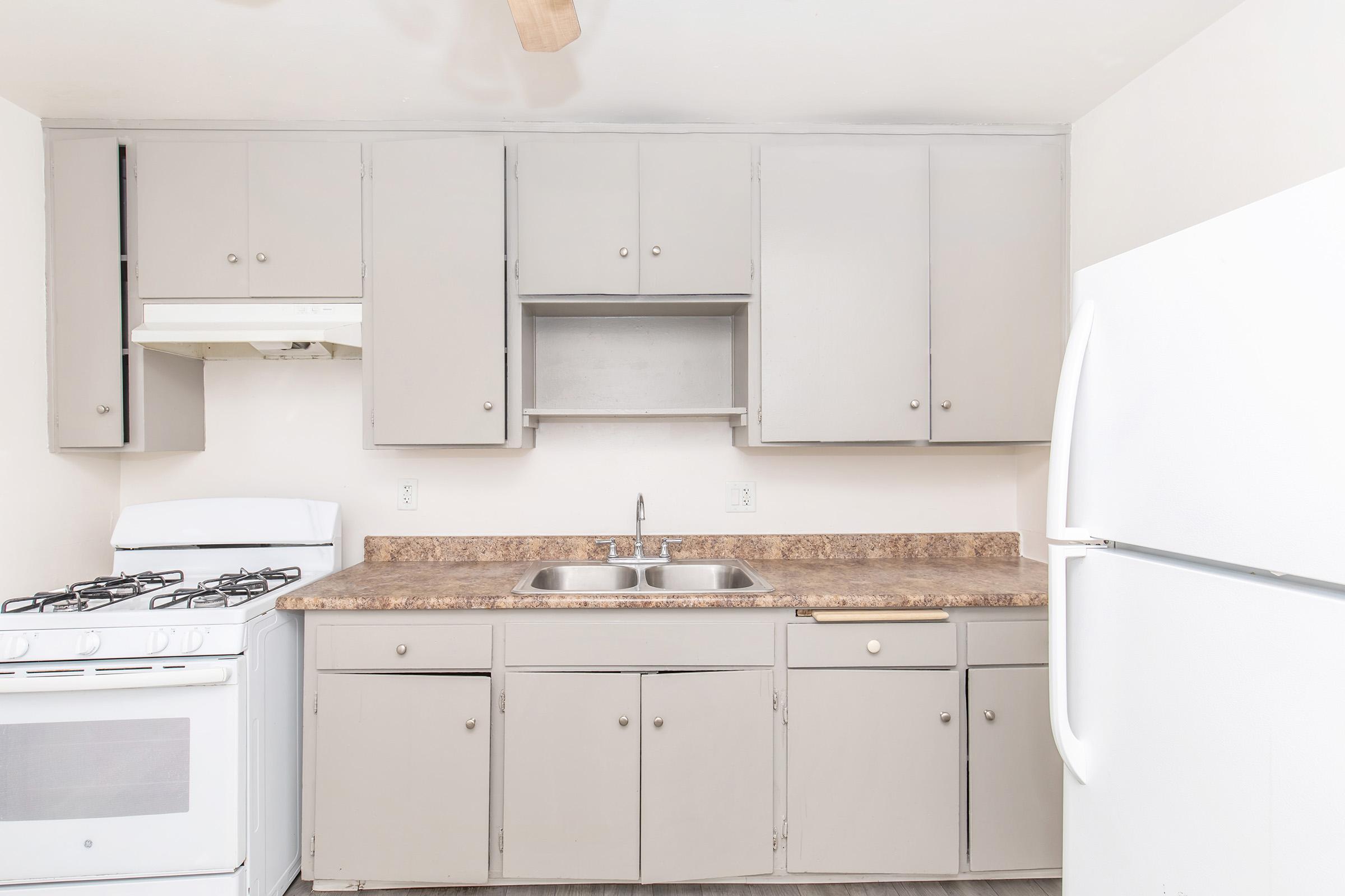 A modern, minimalist kitchen featuring light gray cabinets, a white gas stove, and a double sink with a faucet. A white refrigerator is visible on the right, and the countertop is a darker, textured surface. The walls are painted in a neutral color, giving the space a clean and airy feel.