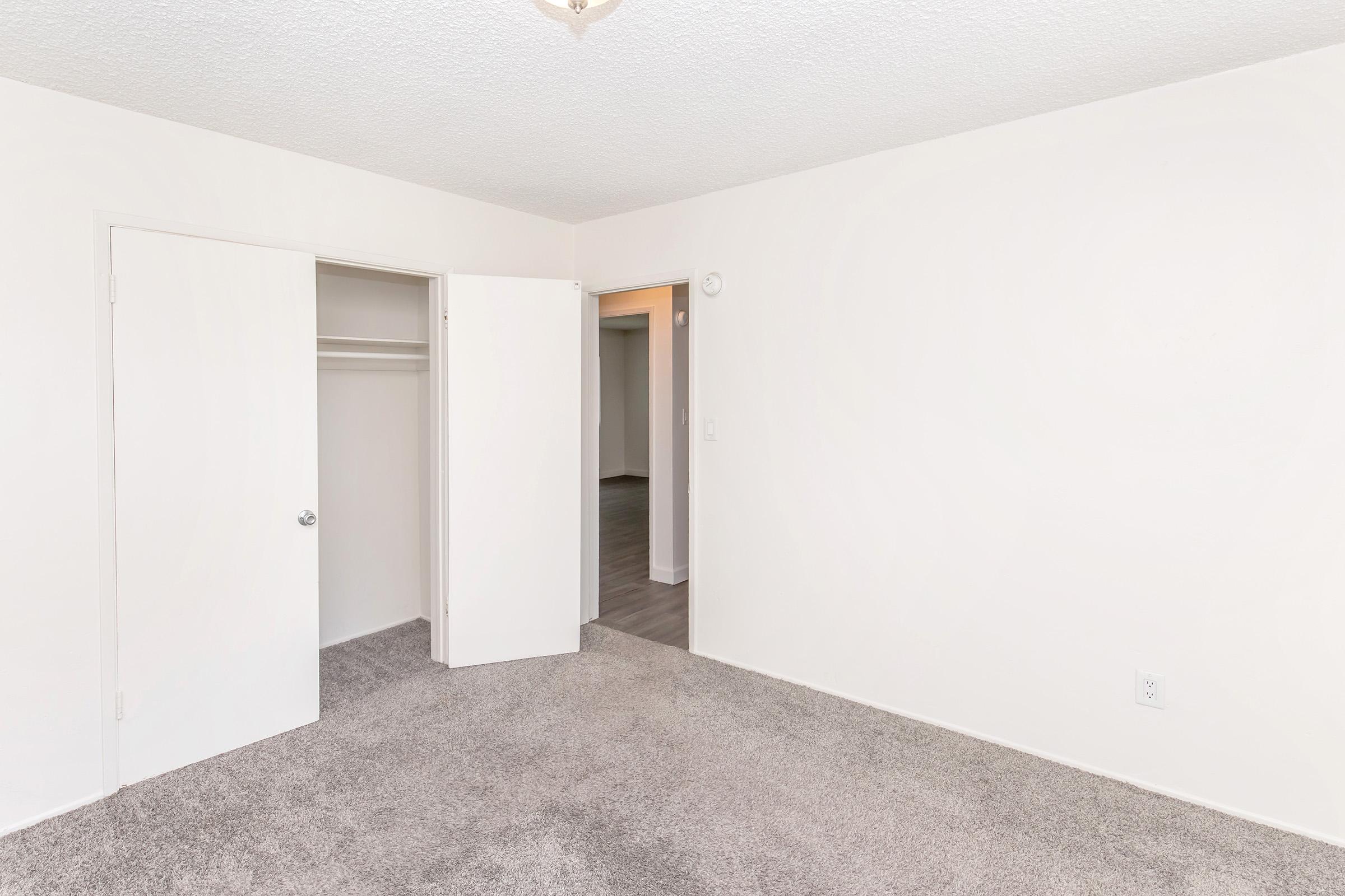 Interior view of a clean, empty room with beige carpet and light-colored walls. A closet with sliding doors is visible on the left, and an open doorway leads to another room on the right. The ceiling features a simple light fixture. The space appears bright and uncluttered, ideal for personalization.
