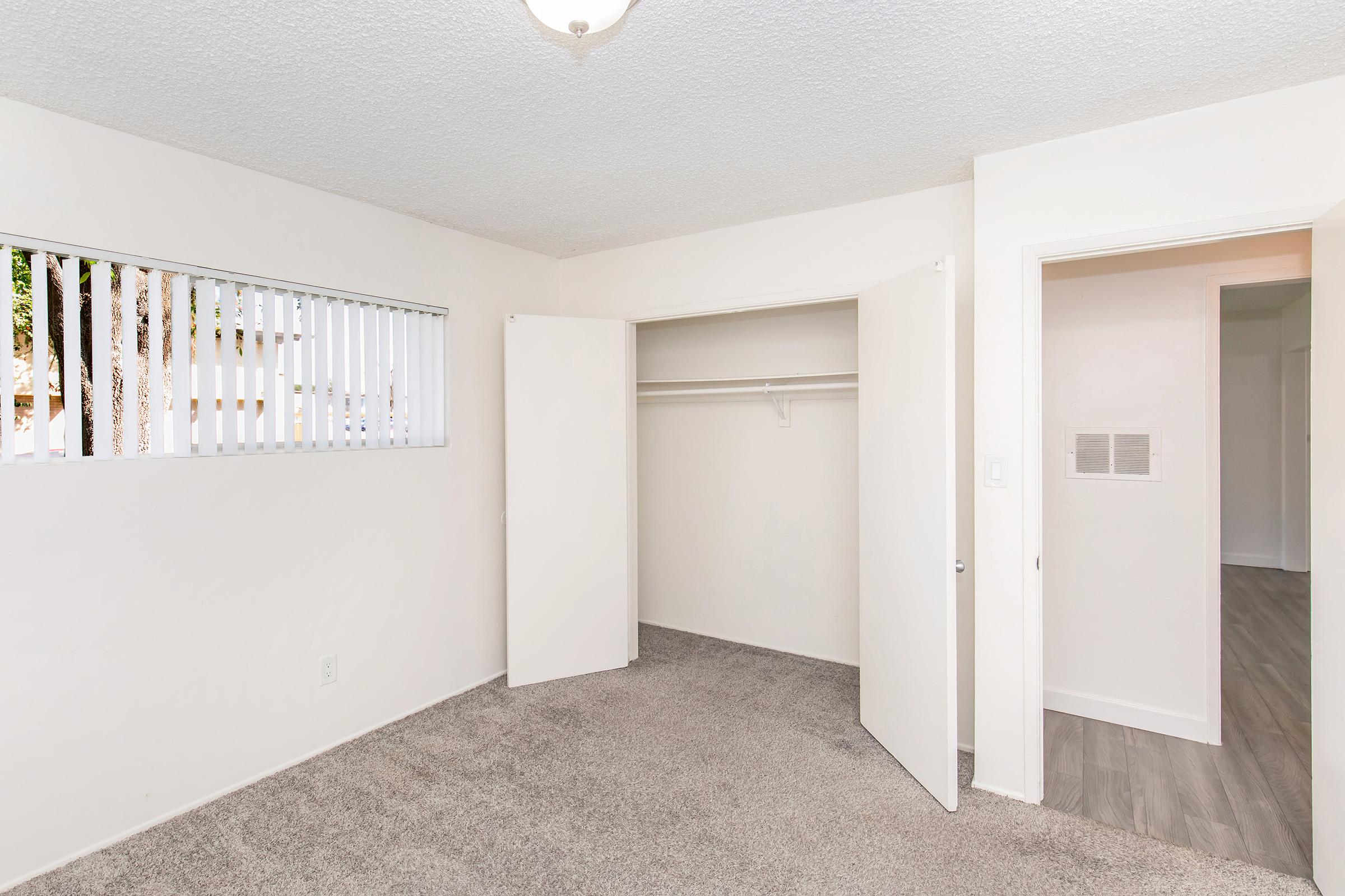 A spacious room featuring light-colored walls and a textured ceiling. There is a closet with sliding doors on one side, and a window with vertical blinds, allowing natural light. The flooring is carpeted, and the room is finished with a light fixture on the ceiling.