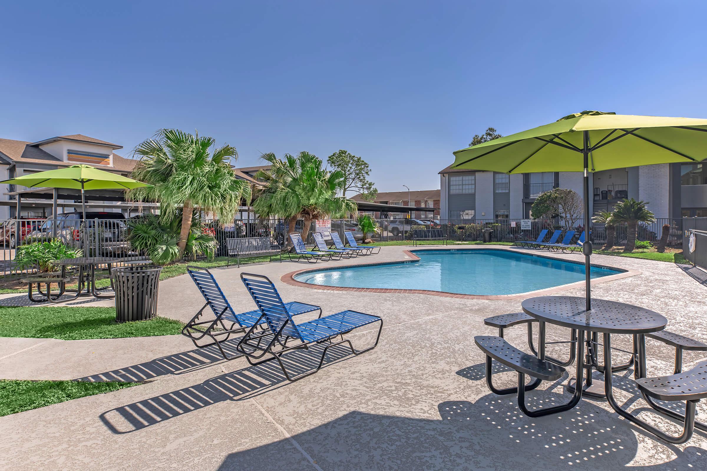 A sunny outdoor pool area featuring a kidney-shaped swimming pool surrounded by lounge chairs and green umbrellas. There are tables for seating, palm trees, and a well-maintained landscape, creating a relaxing atmosphere. Modern apartment buildings are visible in the background.