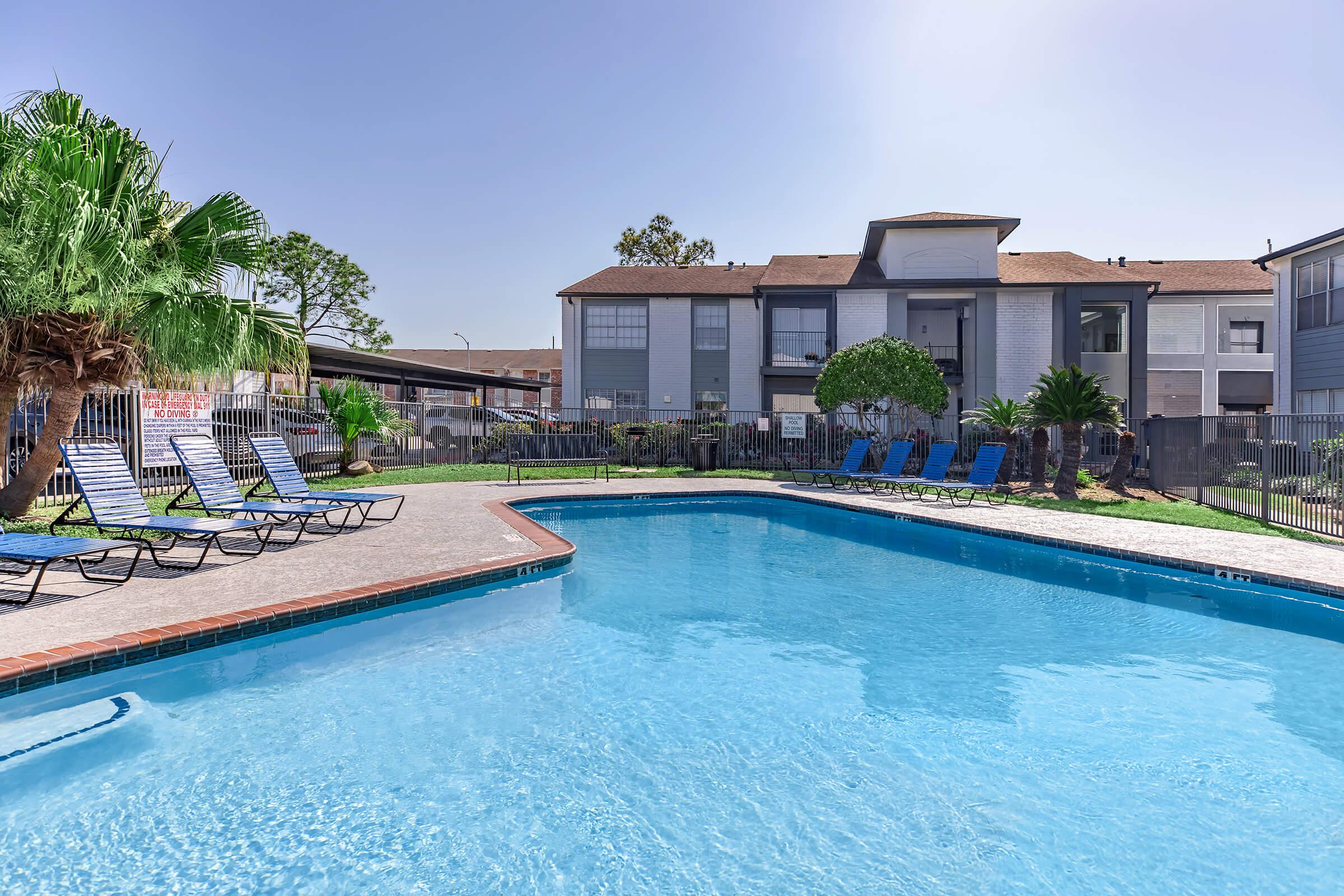 A clear blue swimming pool surrounded by lounge chairs and tropical plants. In the background, there are apartment buildings with balconies. The sky is bright and sunny, creating a relaxing atmosphere.