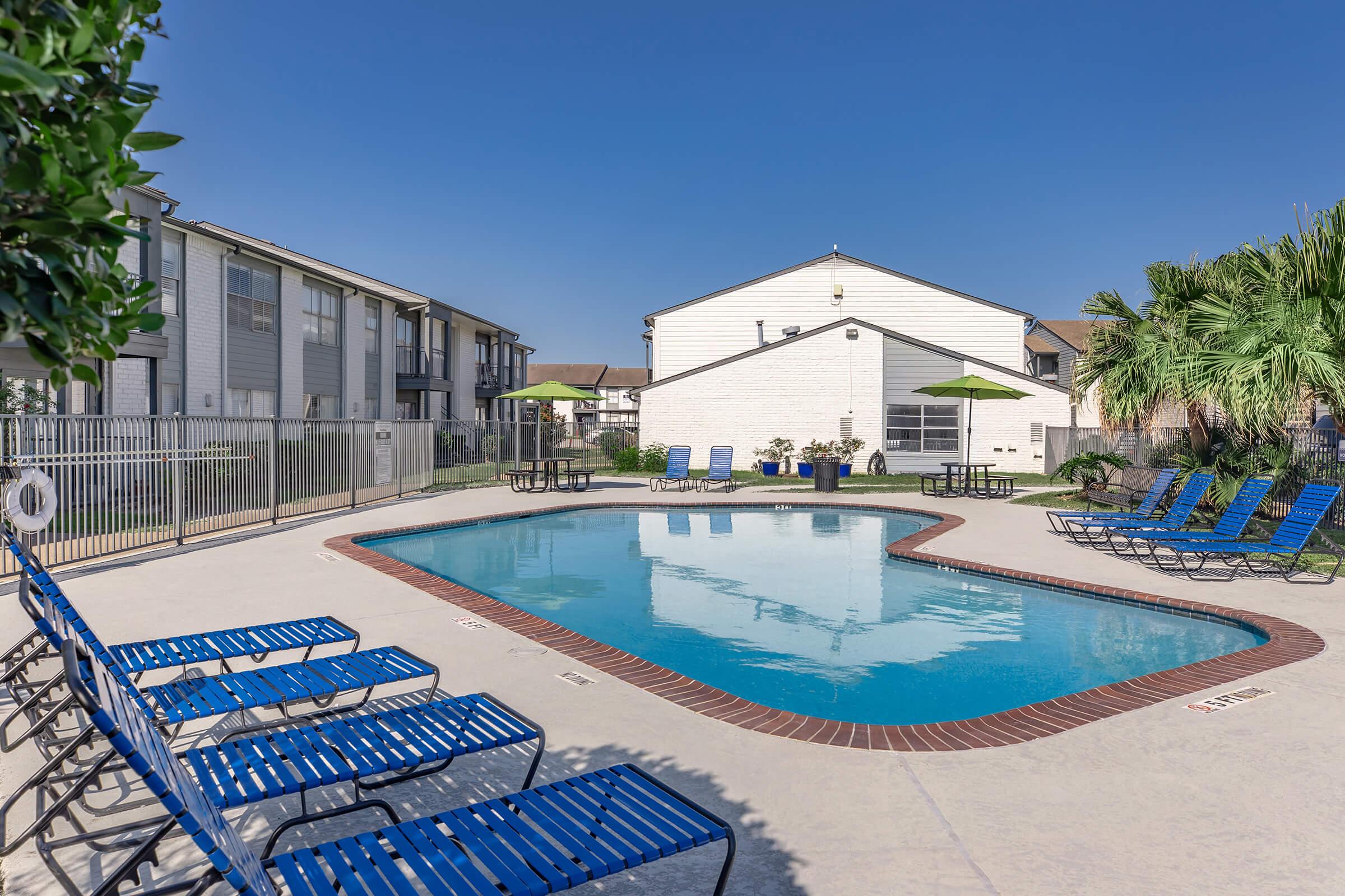 A clear swimming pool surrounded by lounge chairs under a blue sky. The pool area is enclosed with a fence, featuring green umbrellas and tropical plants nearby. In the background, residential buildings can be seen.