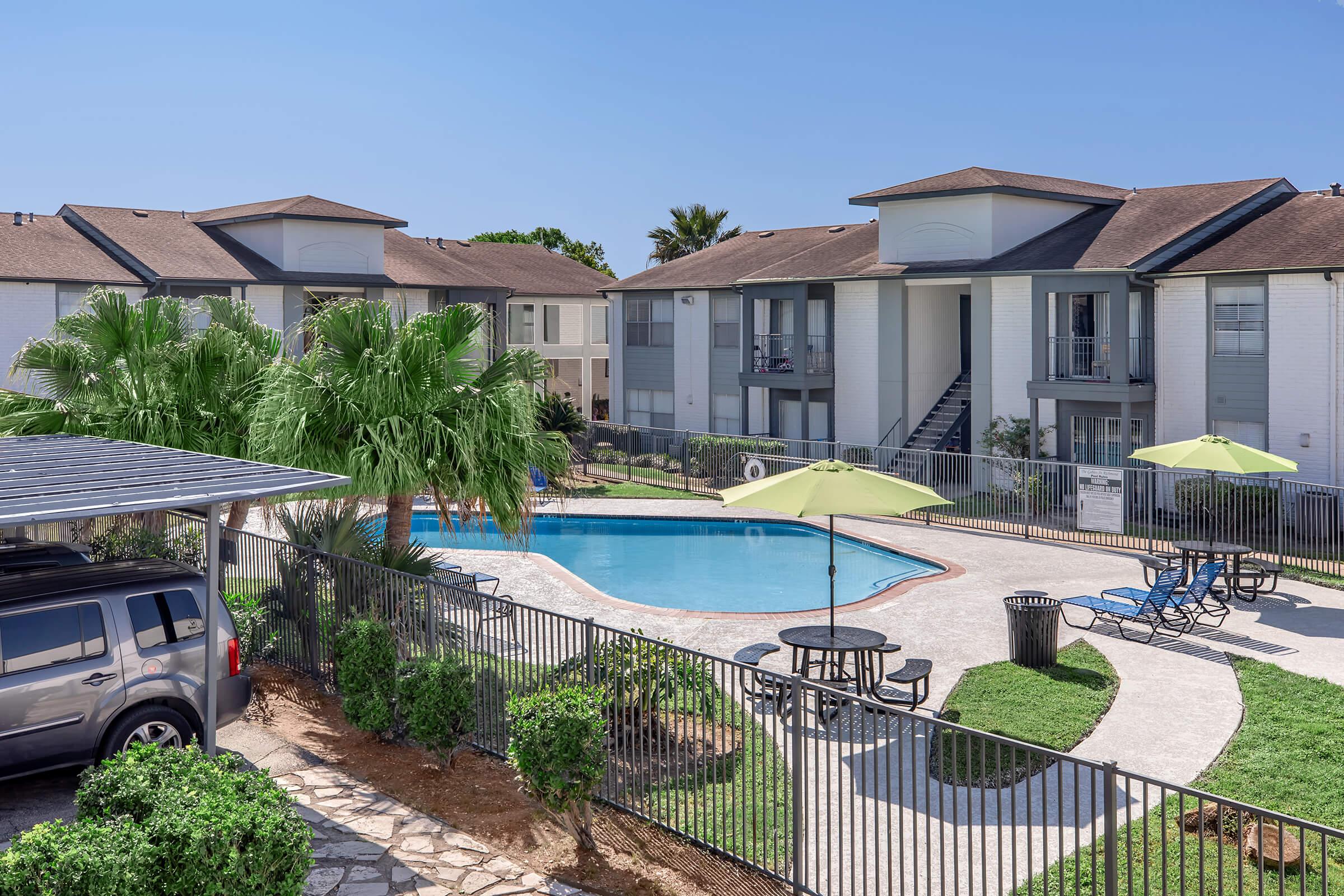View of an apartment complex with a swimming pool. The area features green grass, palm trees, and lounge chairs around the pool. There is a car parked under a shelter and several umbrellas providing shade near the poolside. Clear blue sky above complements the sunny day.