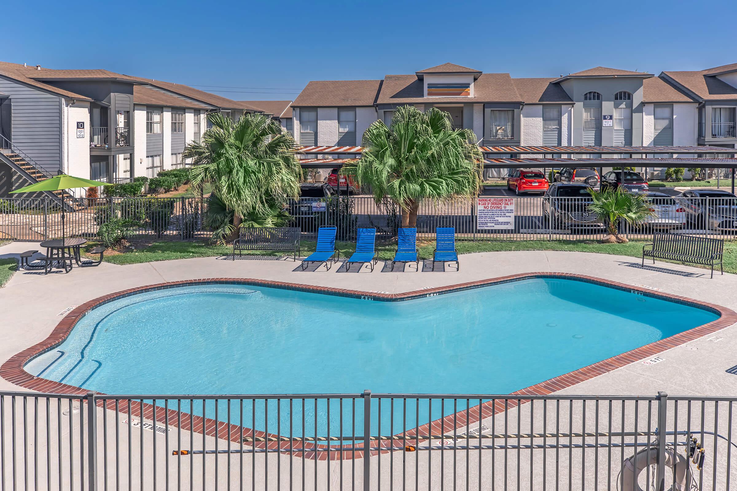 A heart-shaped swimming pool surrounded by lounge chairs and palm trees, with residential buildings in the background. The clear blue sky provides a bright and sunny atmosphere, while cars are parked in the shaded area nearby.