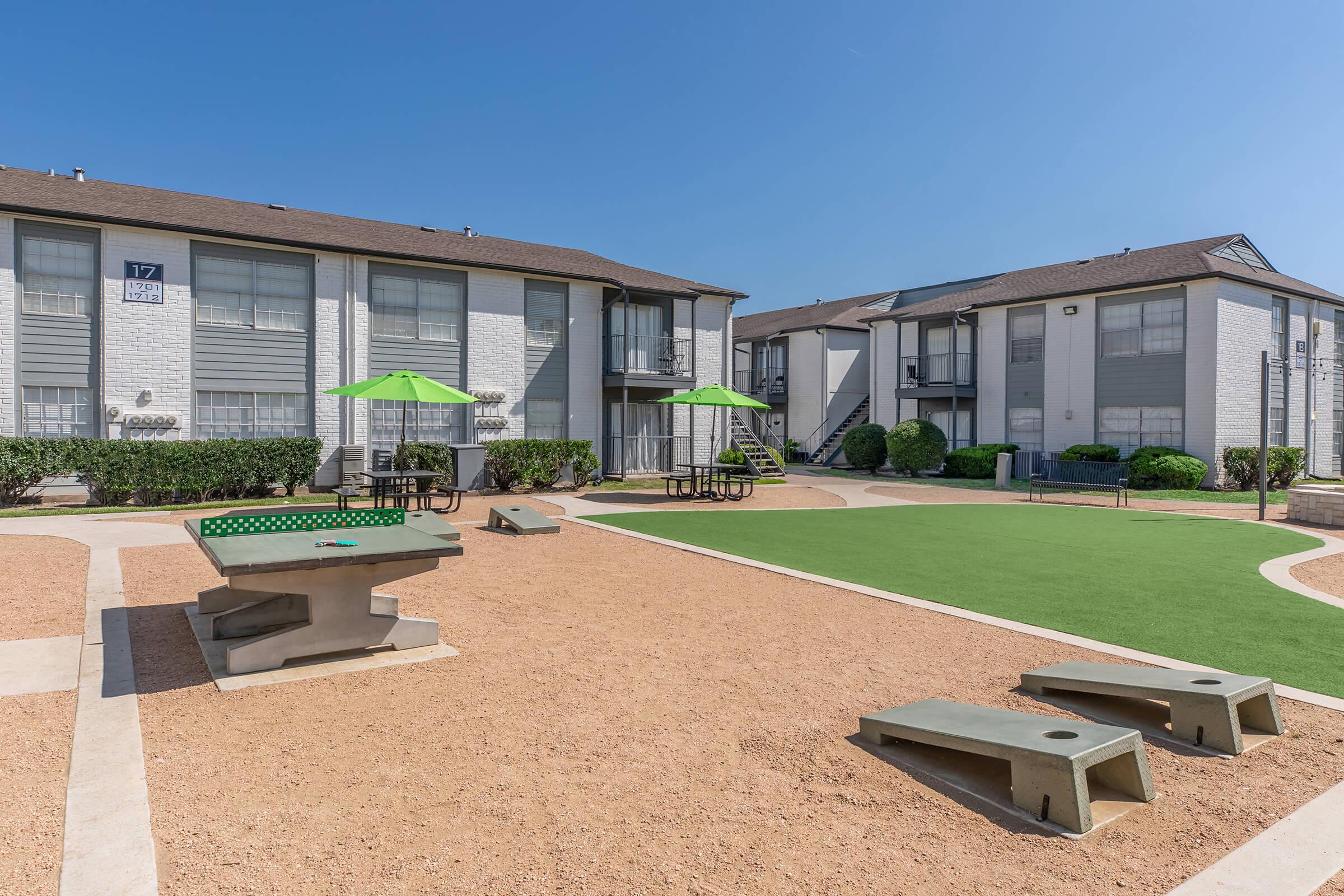 A well-kept outdoor area of an apartment complex featuring a green lawn, picnic tables with green umbrellas, and game tables on a gravel surface. Two buildings are visible in the background under a clear blue sky.