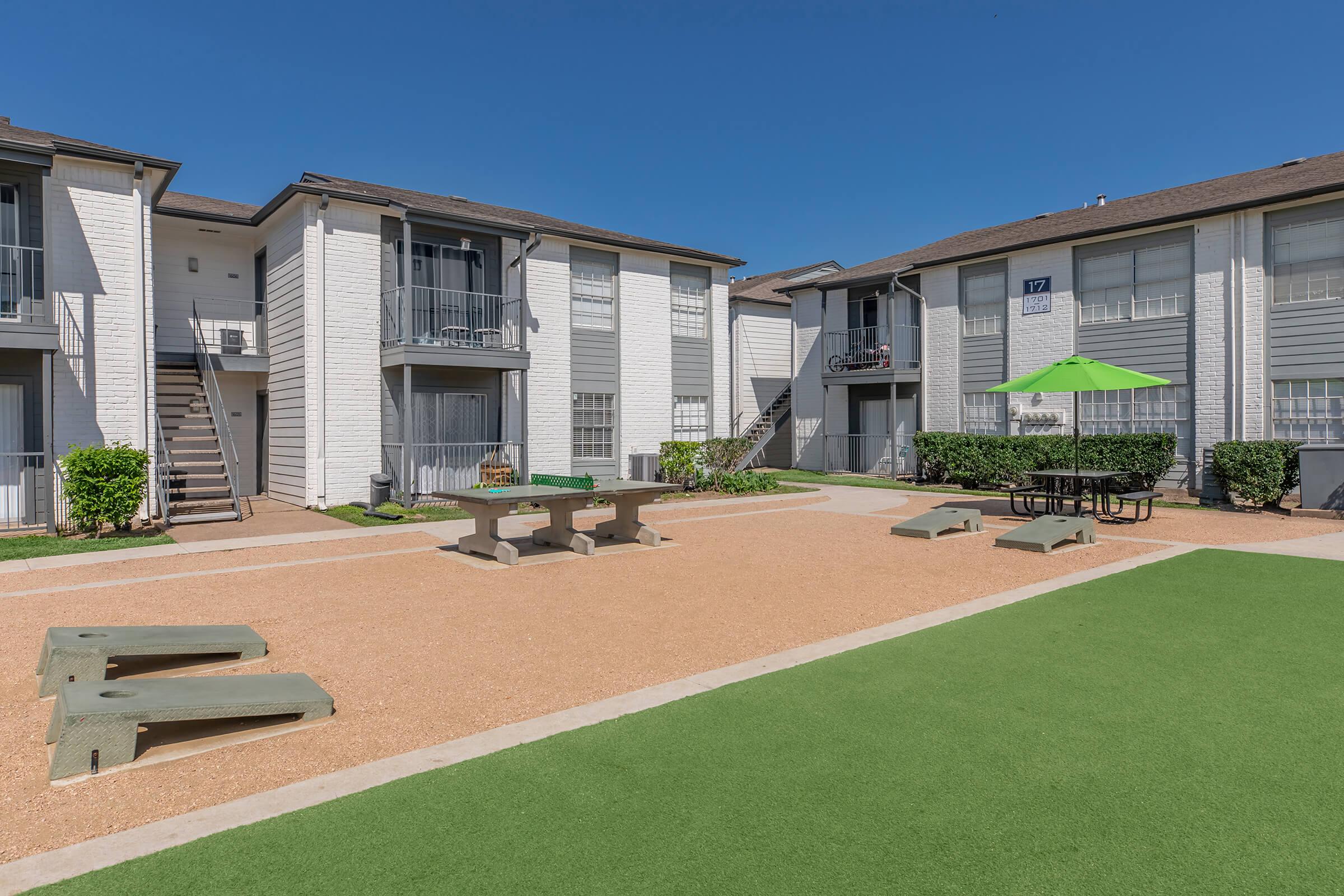 A sunny courtyard area of an apartment complex featuring two buildings, green grass, picnic tables, and a green umbrella. The space is designed for outdoor activities and relaxation, with a clear blue sky overhead.