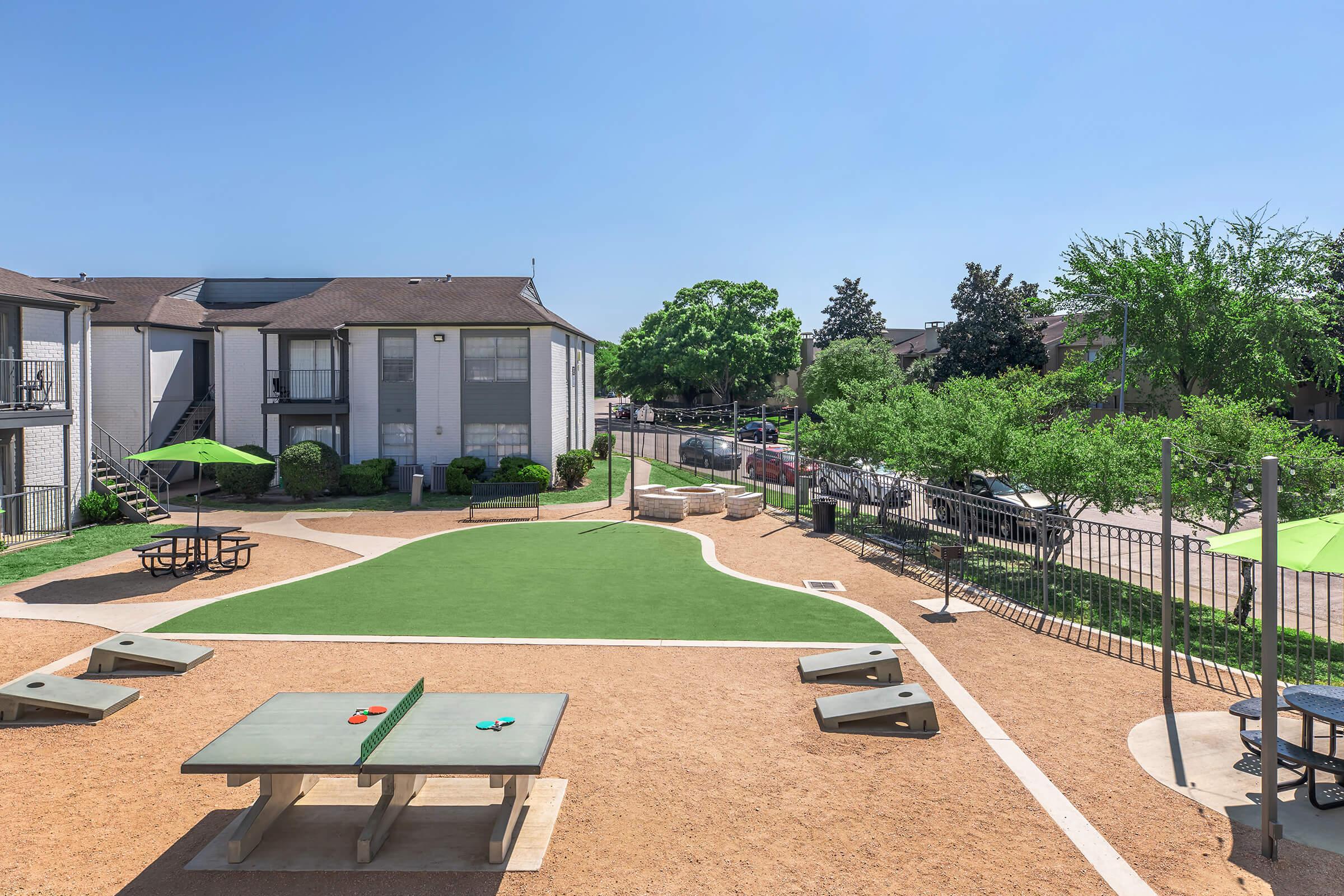 View of a landscaped outdoor area featuring green grass, picnic tables, and game tables set up for activities. Surrounding trees and shrubs provide greenery, and there are buildings in the background with clear blue skies above. The space is designed for recreational use and social gatherings.