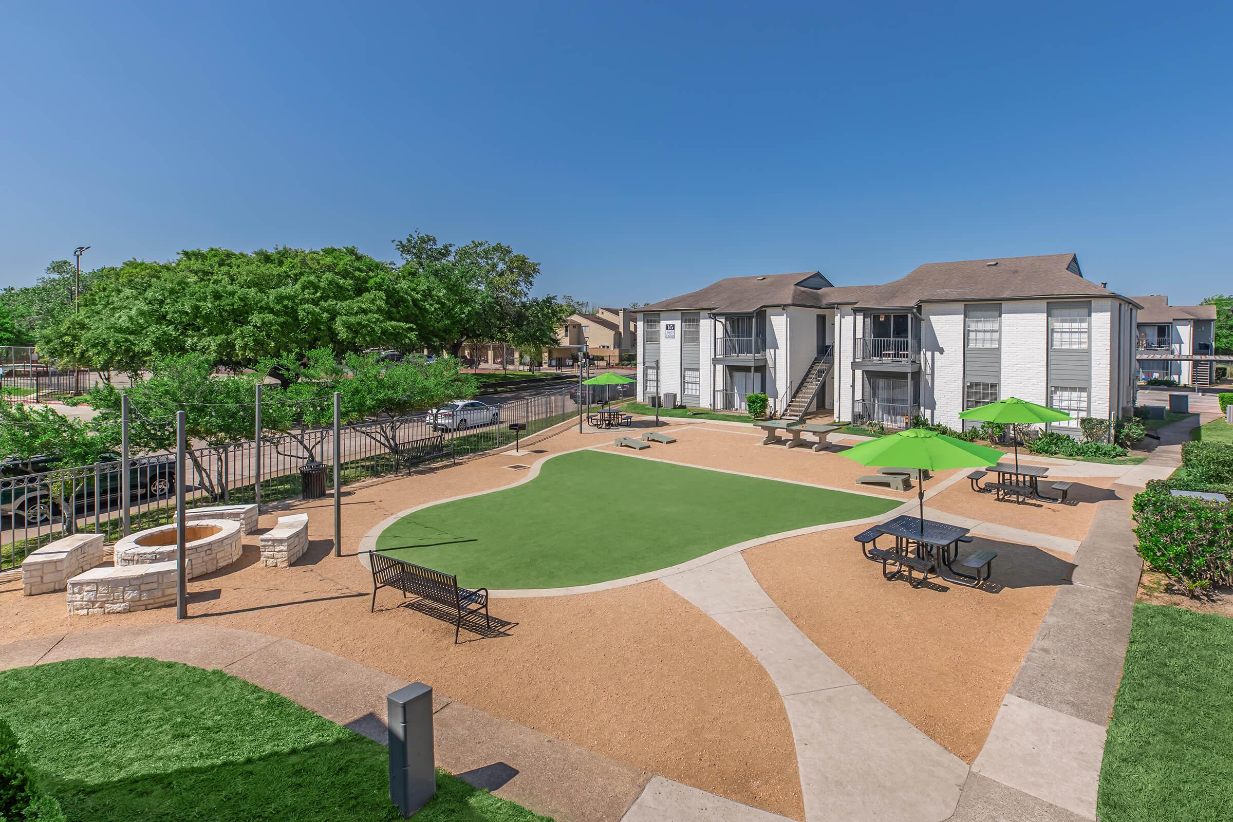 A well-maintained outdoor area featuring a green lawn, decorative stone seating, picnic tables with green umbrellas, and a walking path. Surrounding the space are landscaped bushes and trees, with a multi-story residential building in the background under a clear blue sky.