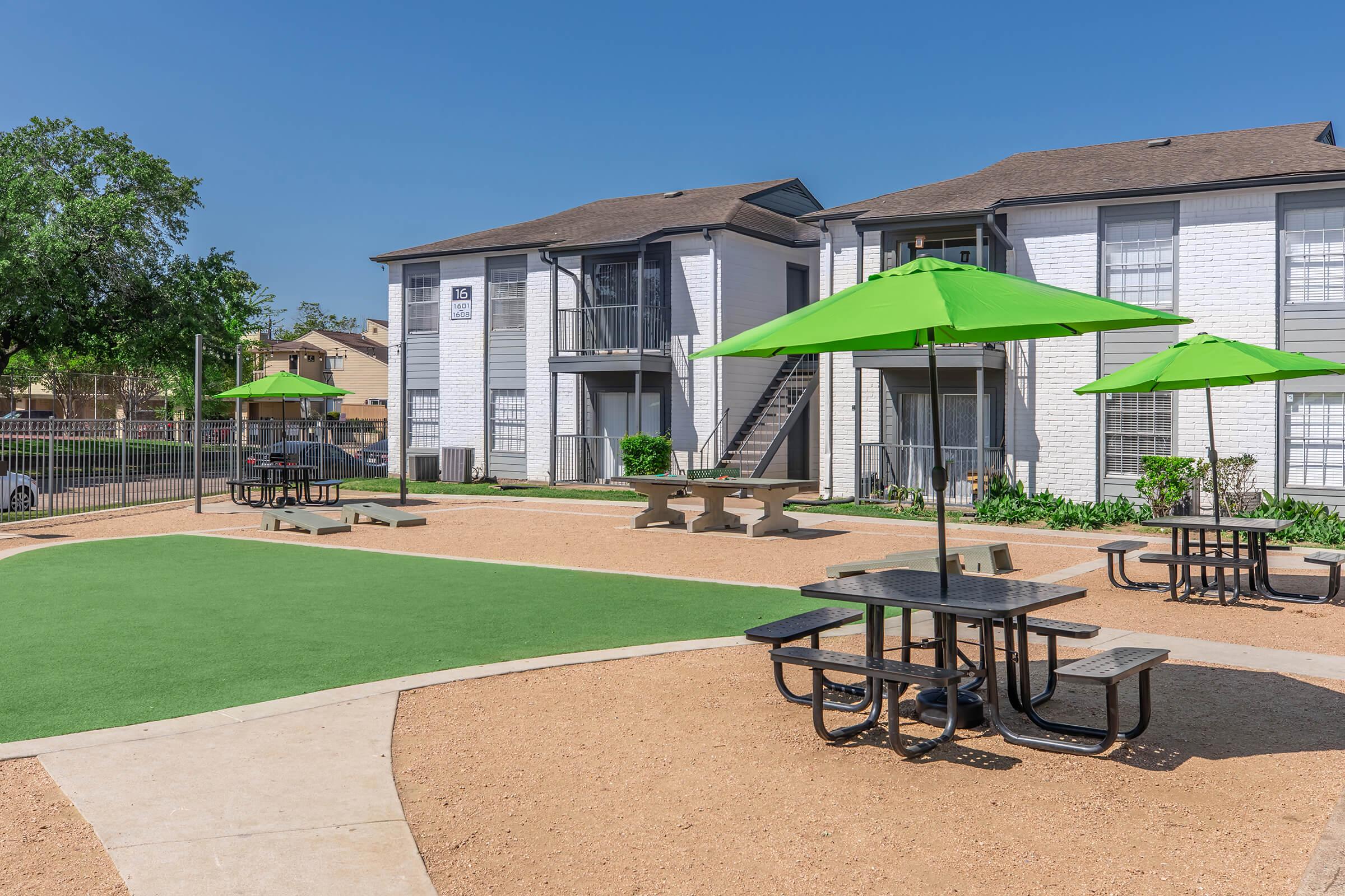 Outdoor area of an apartment complex featuring several picnic tables with attached seating, green umbrellas, and a well-maintained lawn. The buildings in the background are two-storied, with balconies and a clear blue sky above. The setting appears inviting and suitable for gatherings.