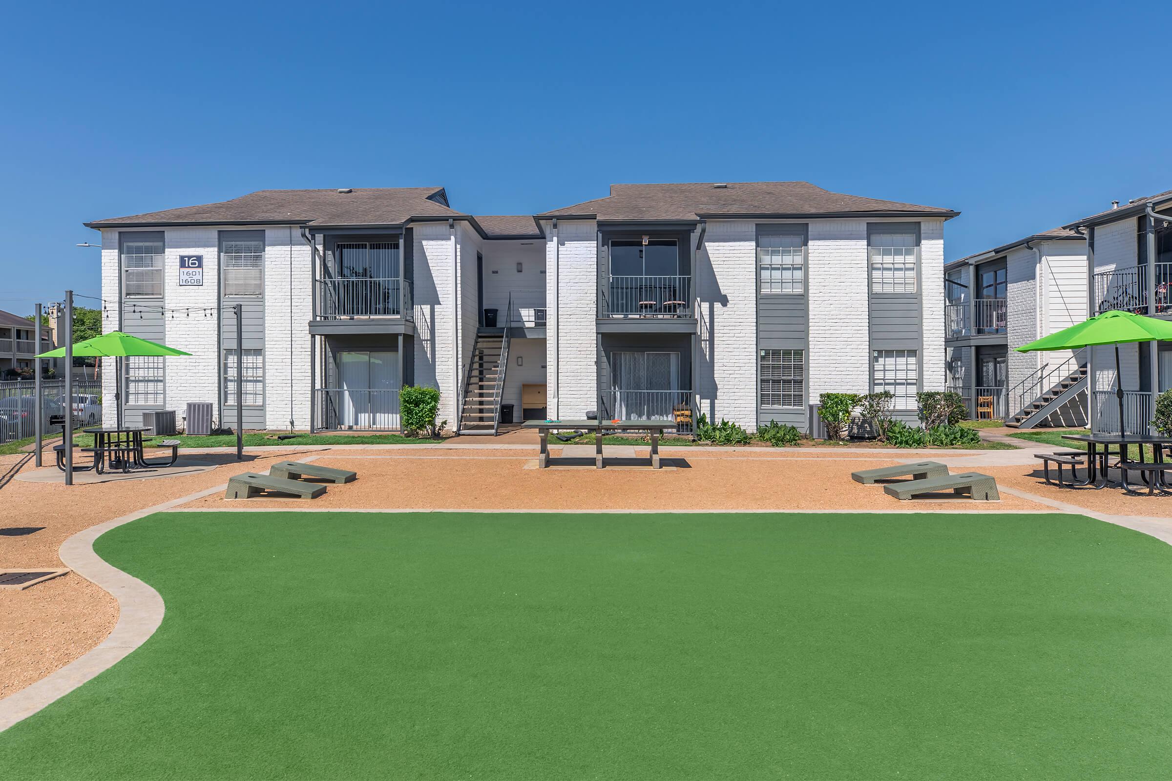 Courtyard view of a residential apartment complex featuring two-story buildings, green lawns, picnic tables, and umbrellas. The sky is clear and blue, creating a bright and inviting atmosphere.