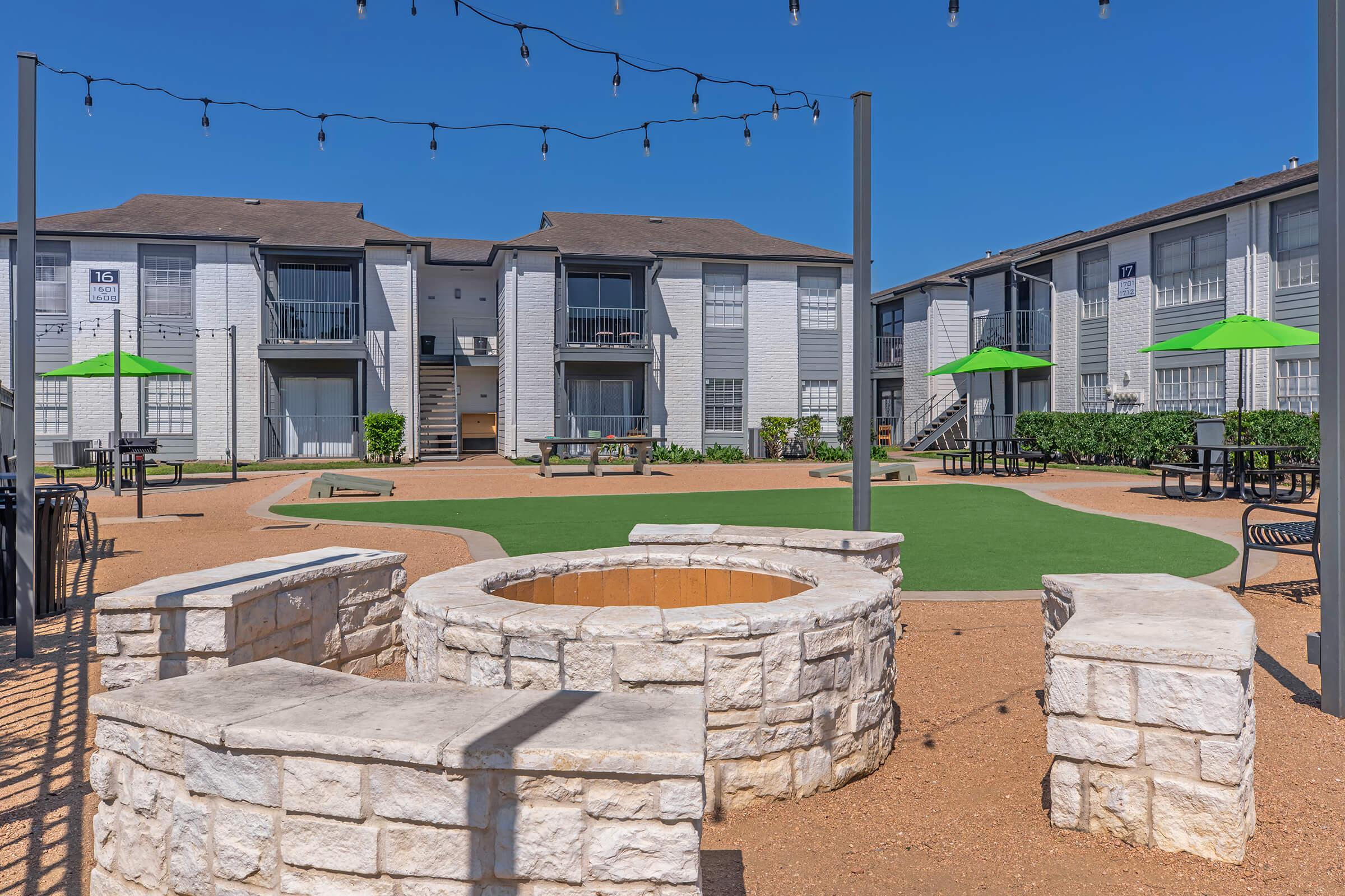 Courtyard of an apartment complex featuring a stone fire pit, green grass area, and outdoor seating. Surrounding the space are light string decorations and several green umbrellas over tables. The buildings are modern with multiple windows, creating a welcoming community atmosphere.