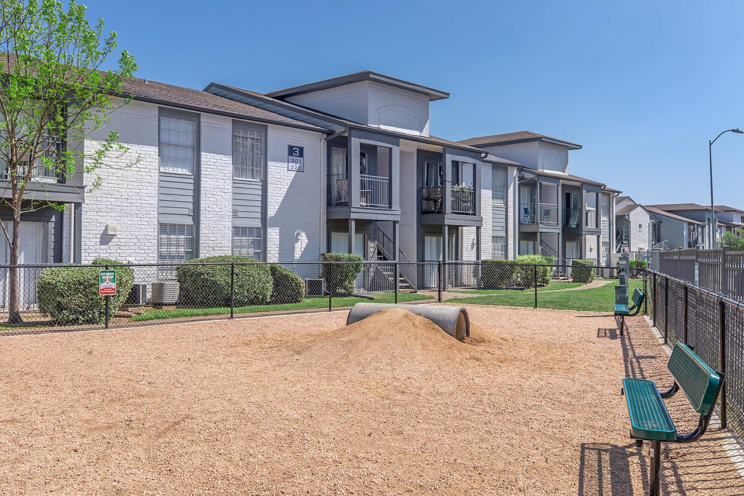 A view of a residential apartment complex featuring two-story buildings with balconies. In the foreground, a fenced play area with a mound of sand and a metal tunnel. Green benches are placed along the perimeter, surrounded by manicured lawns and shrubbery, under a clear blue sky.