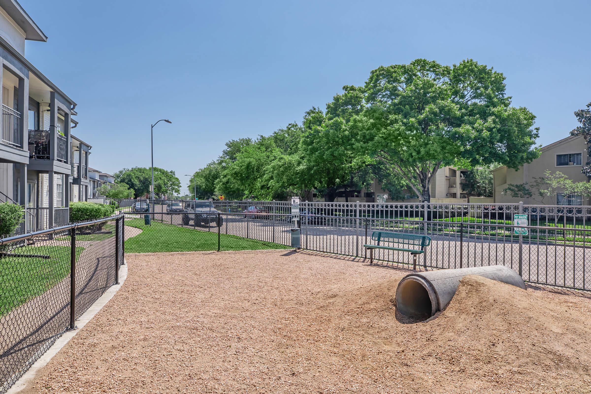 A fenced playground area with a sand-covered surface, featuring a large drainage pipe, a bench, and green grass. Surrounding the playground are residential buildings with trees visible in the background, under a clear blue sky.