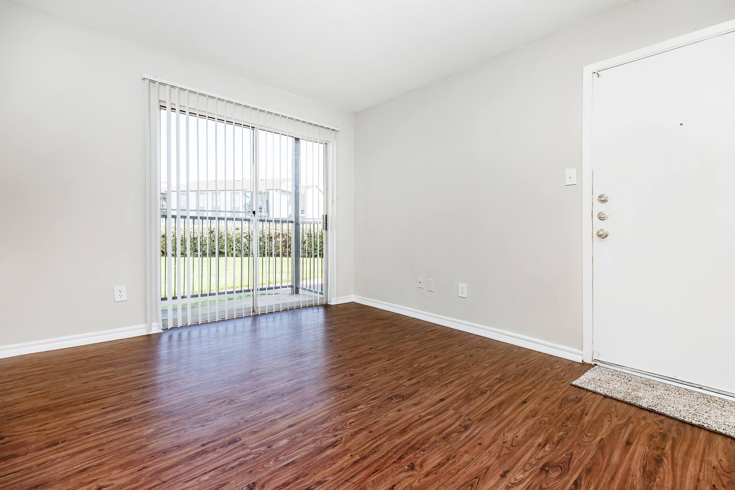A bright and empty room with light-colored walls and wood laminate flooring. A sliding glass door with vertical blinds leads to a patio area with a view of greenery outside. The door to the room is visible on the right, and the space looks clean and ready for furniture.