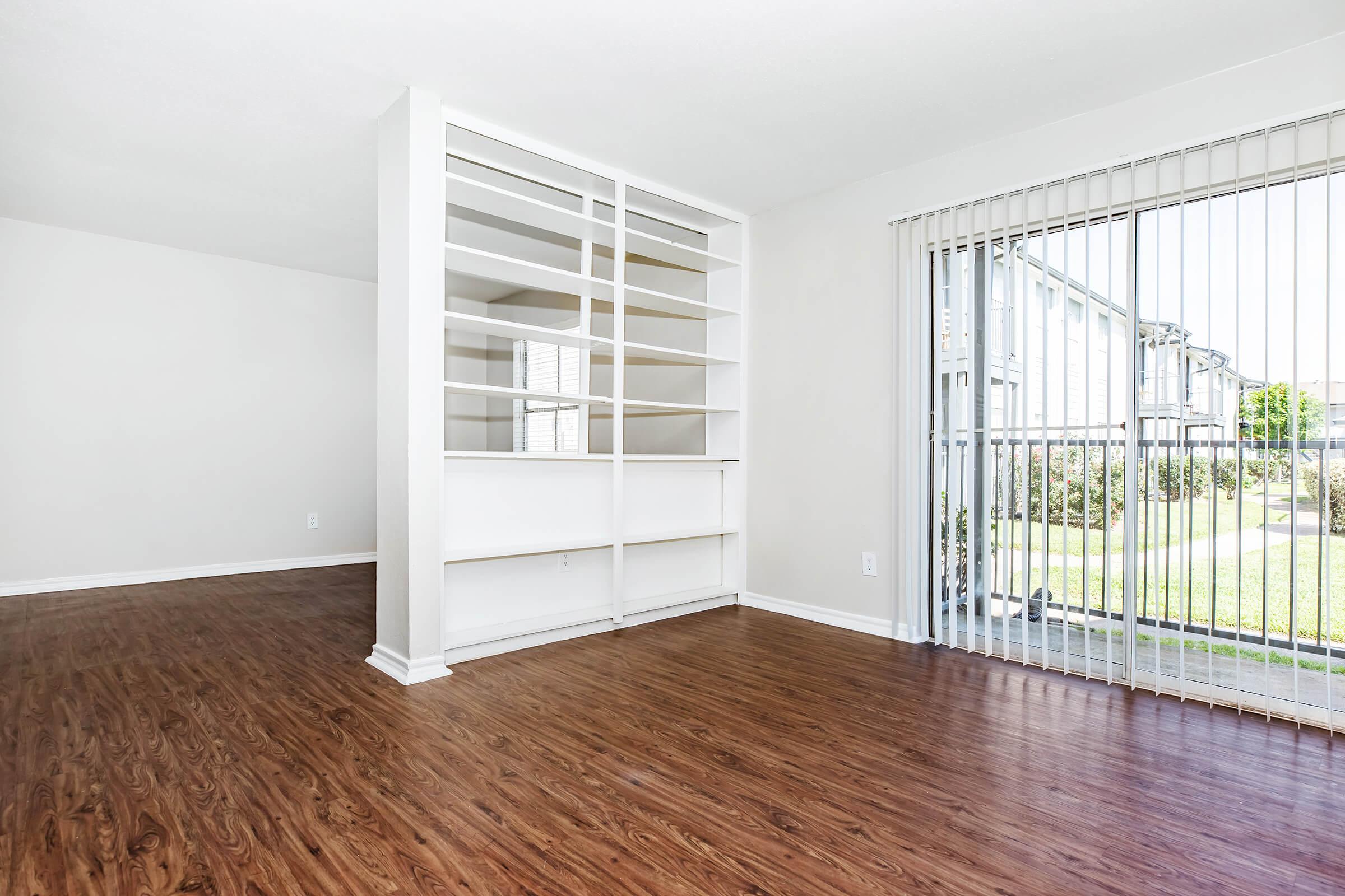 A bright, empty room featuring laminate wood flooring and a white shelving unit dividing the space. Large sliding glass doors lead to an outdoor area with greenery. The walls are painted in a light color, creating an open and airy atmosphere.