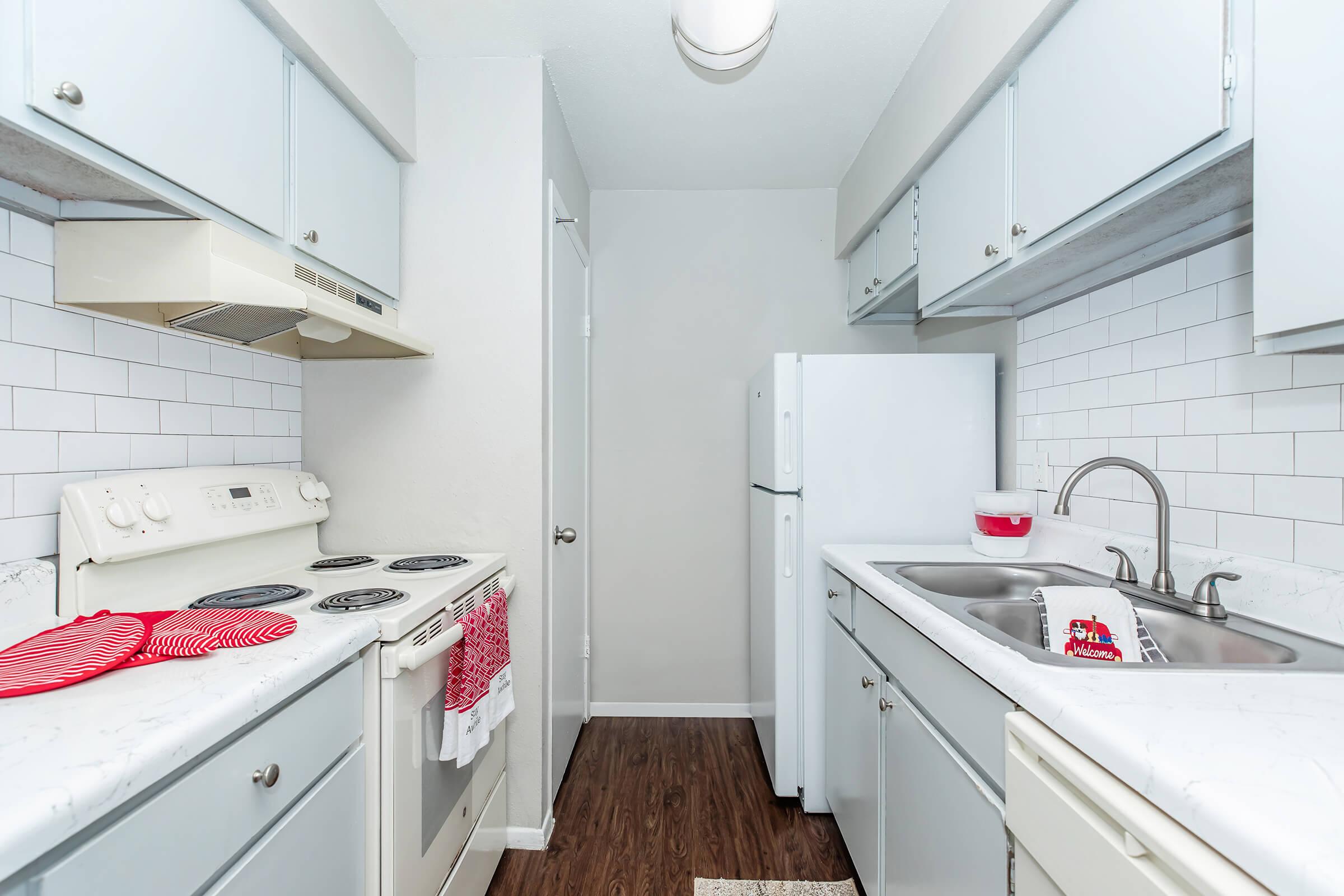 A modern kitchen featuring light blue cabinets, a white countertop, and stainless steel appliances. The layout includes a stove with an overhead range hood, a double sink, and a refrigerator. The walls are adorned with white subway tiles, and the floor is a dark wood finish, creating a bright and inviting space.