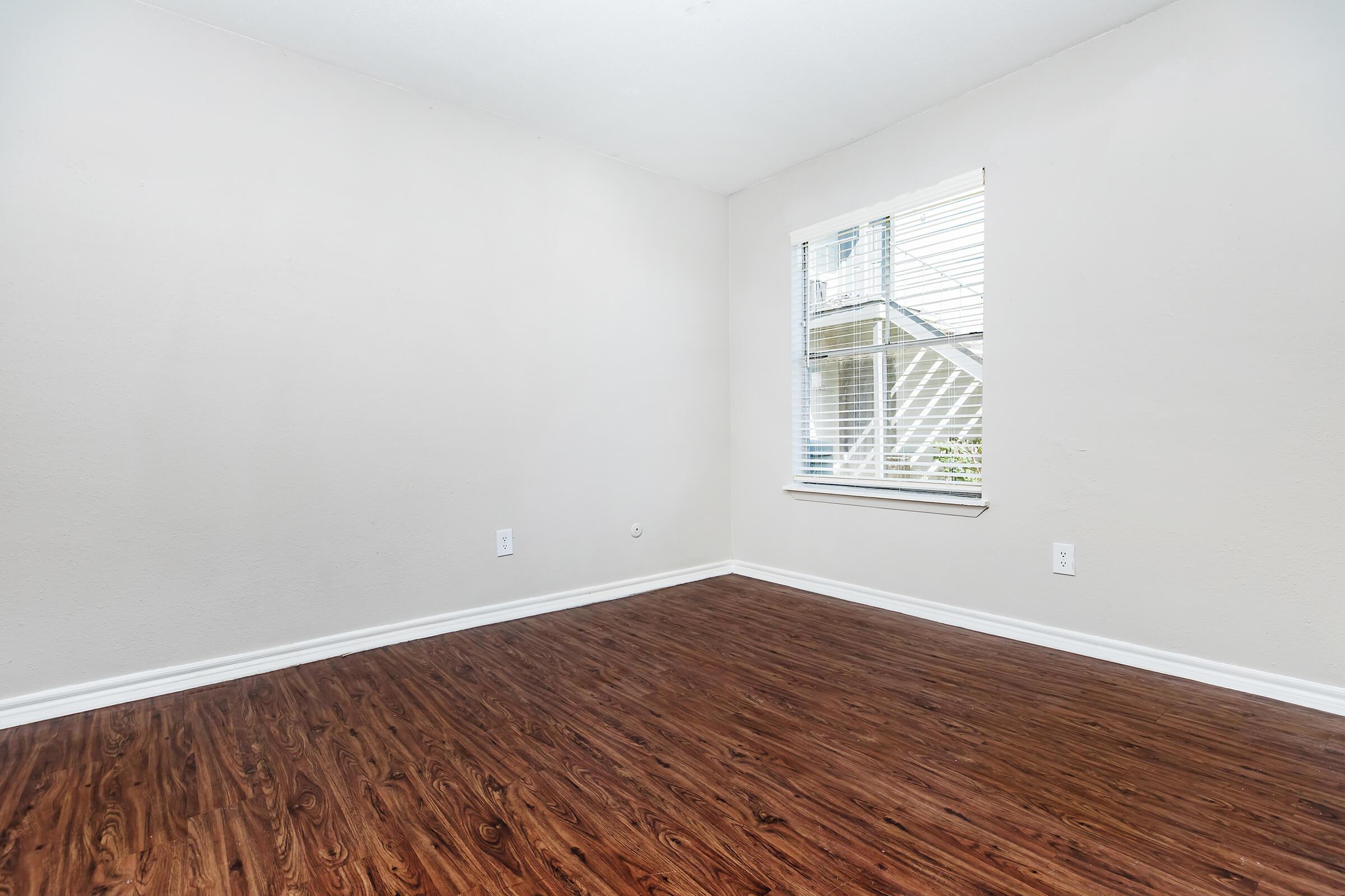 A spacious empty room with light-colored walls and brown laminate flooring. There is a window with blinds allowing natural light to enter, but no furniture or decor present, creating a minimalist and clean atmosphere.