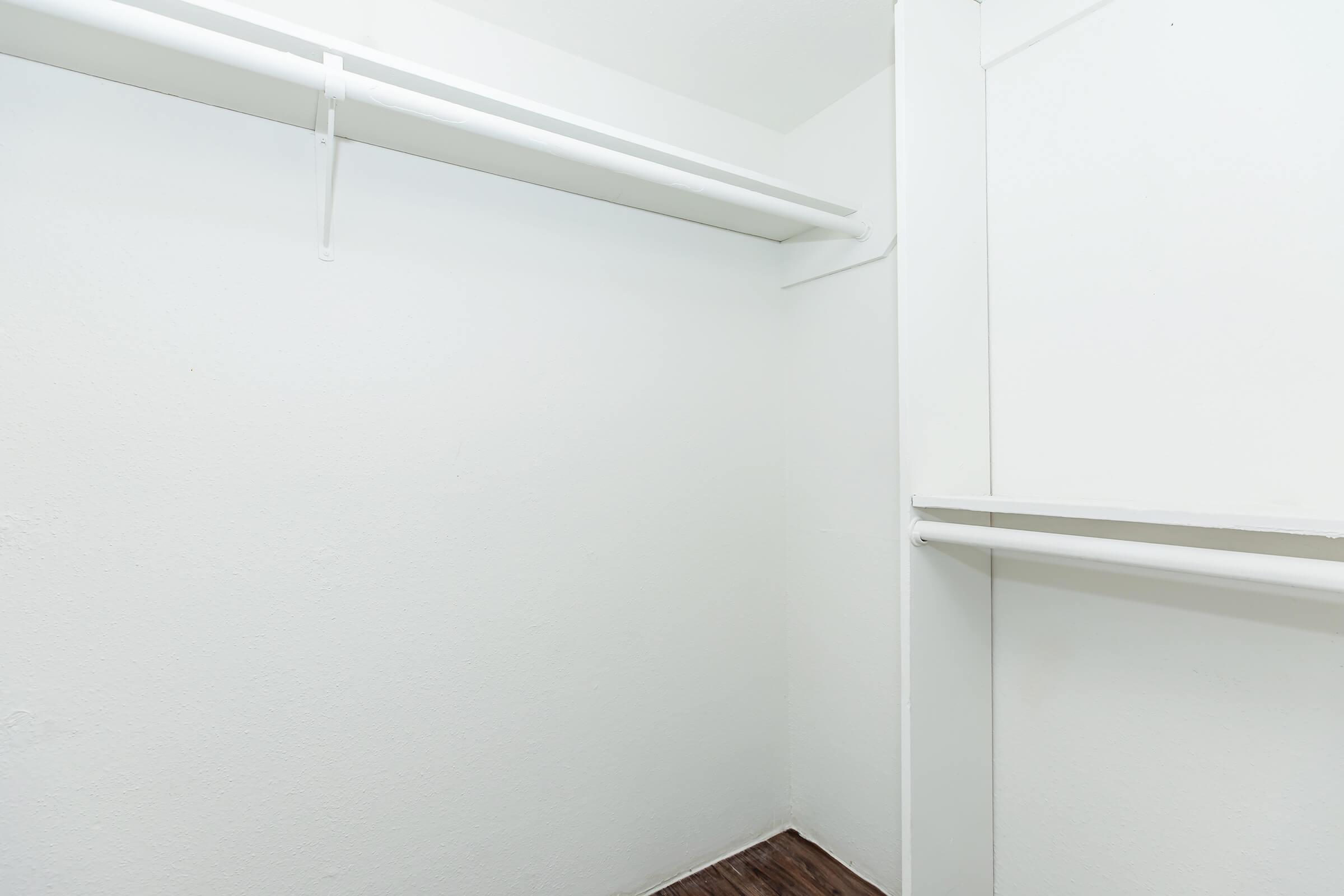 Empty closet with white walls and a wooden floor. Two horizontal clothing rods are mounted on the walls, offering space for hanging clothes. The overall appearance is minimalist and clean, with no additional items or furnishings present.