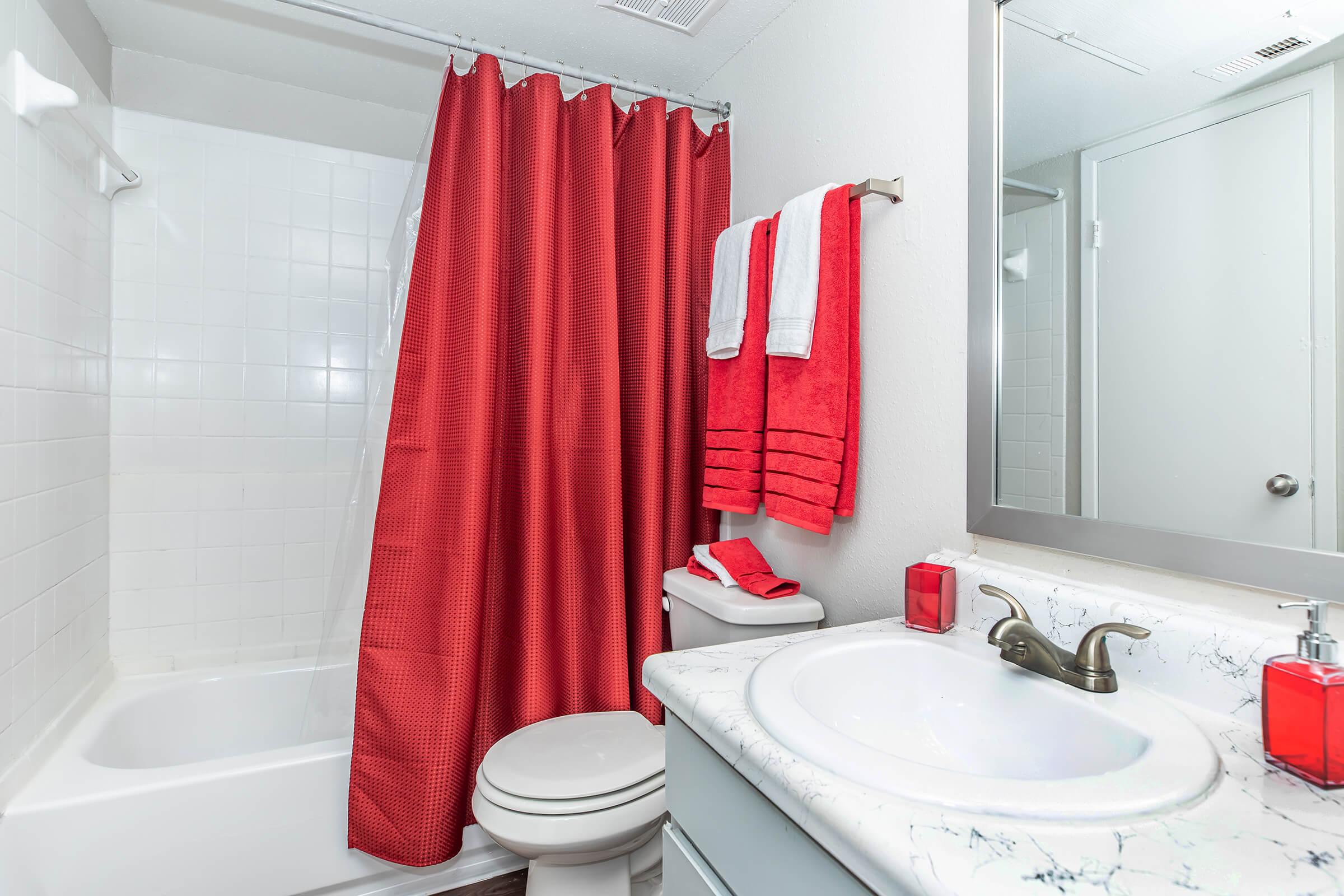A modern bathroom featuring a white bathtub and shower combination, a red shower curtain, red towels hanging on a rack, a white toilet, and a marble countertop sink with a soap dispenser. The walls are light-colored, creating a bright and clean atmosphere.