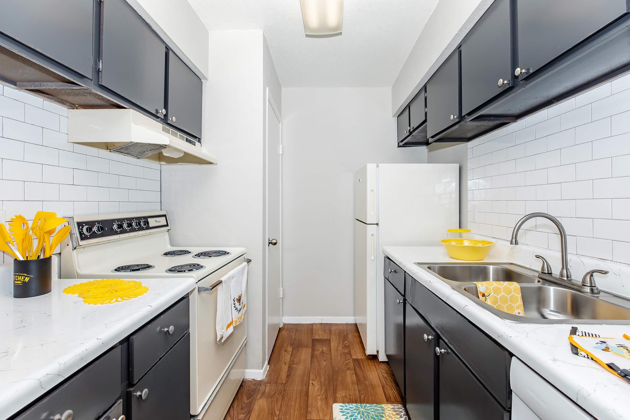 A modern kitchen featuring dark cabinetry, stainless steel appliances, and a white tile backsplash. The countertops are light-colored and decorated with yellow utensils and a bowl. Natural light illuminates the space, which has a laminate wood floor and a clean, organized look.
