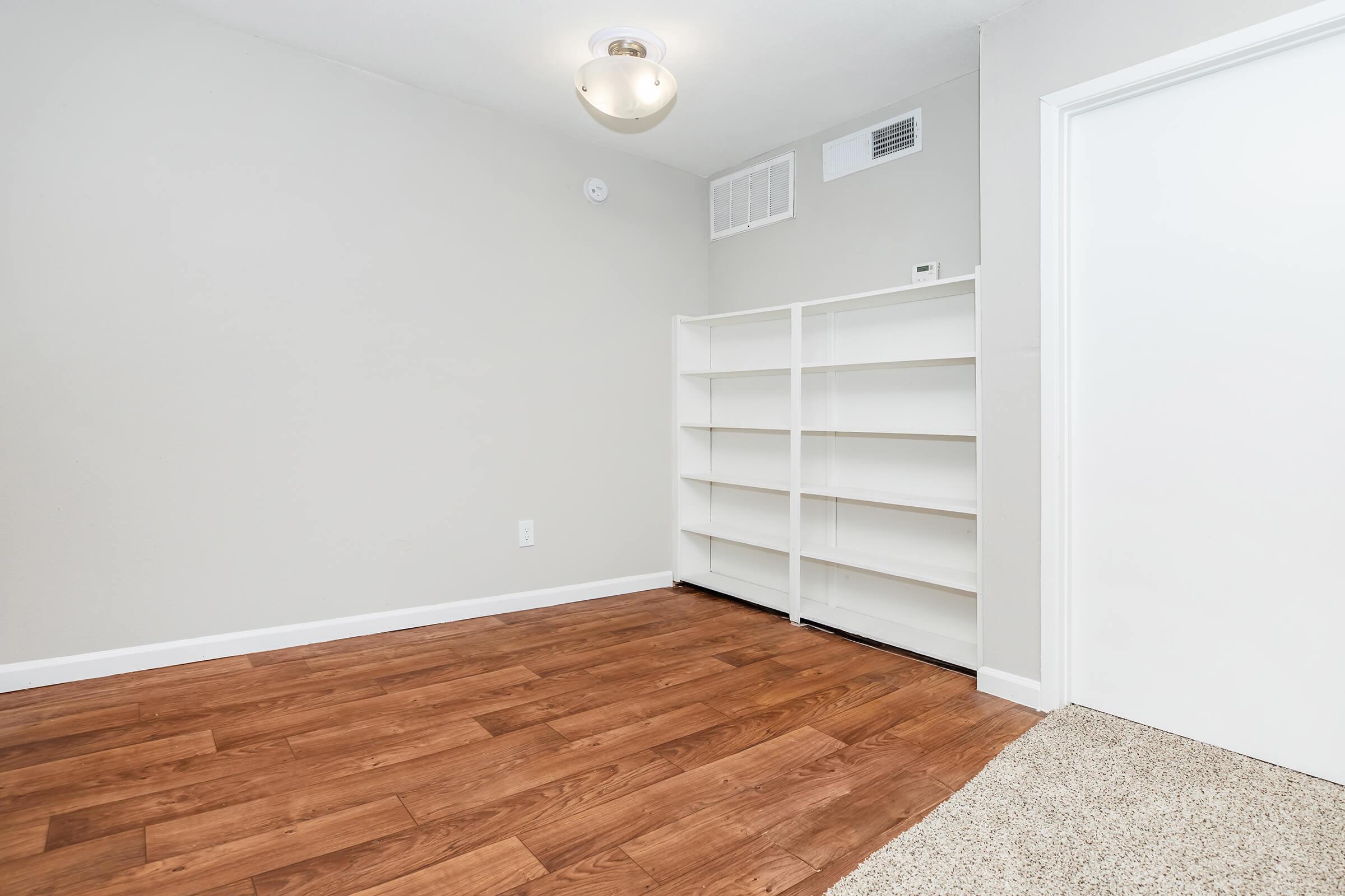 An empty room with light-colored walls and wooden flooring. There is a ceiling light fixture, an air vent, and a white bookshelf along one wall. A carpeted area is visible near the entrance, and the room has a simple, minimalistic design.