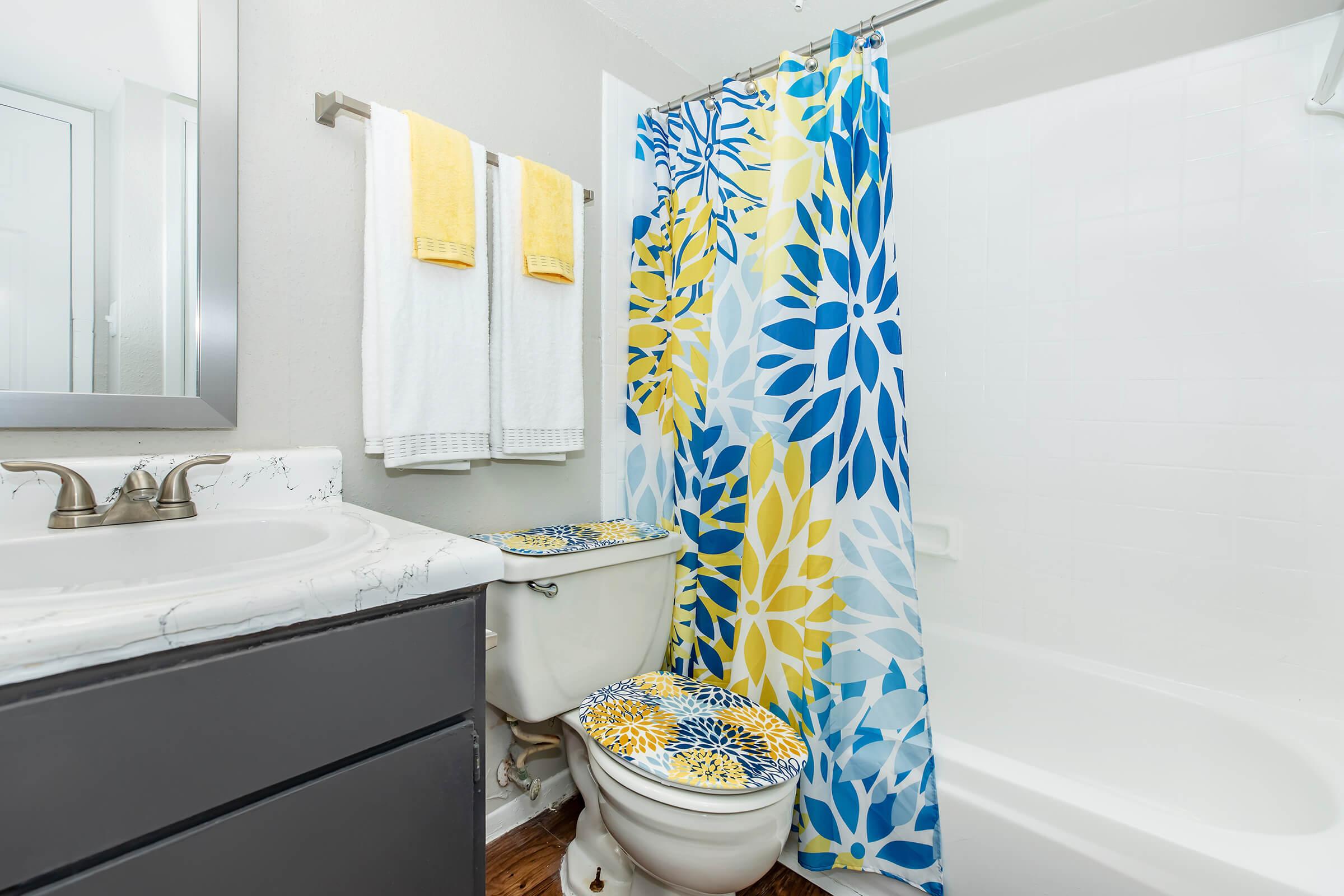 A bright and modern bathroom featuring a bathtub with a shower, a decorative shower curtain in blue and yellow floral patterns, and matching toilet seat covers. The sink area includes a marble countertop, a mirror, and neatly arranged white towels on a towel rack.