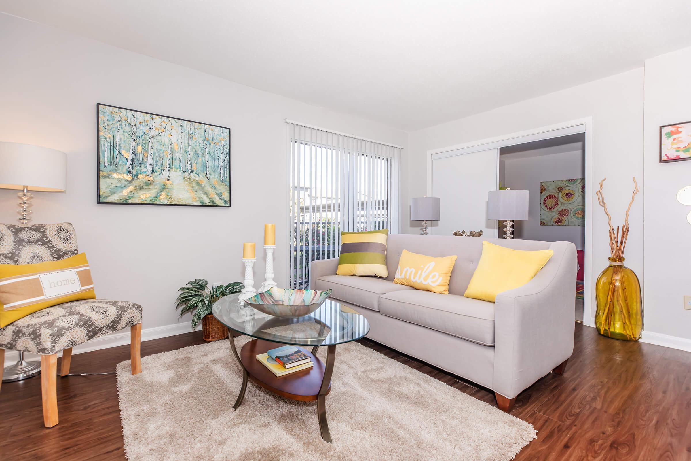 A cozy living room featuring a light gray sofa with yellow and white pillows, a round glass coffee table, and a stylish armchair. There is a colorful artwork on the wall, wooden flooring, and natural light coming through the windows. Decorative items include a bowl and a vase on the table.