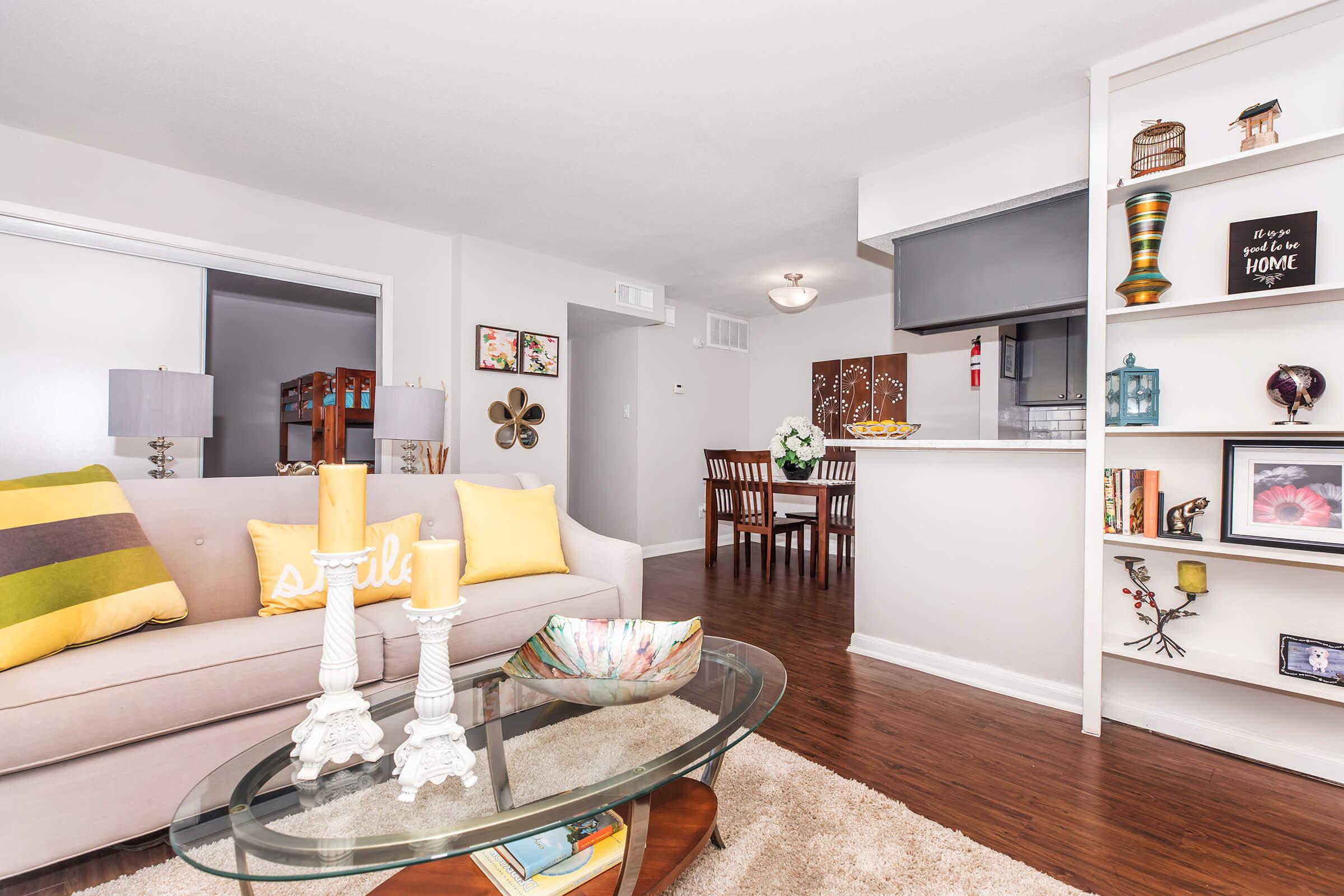 A modern, well-decorated living room featuring a beige sofa with colorful cushions, a glass coffee table with candles, and a cozy rug. In the background, there's a dining area with a wooden table and chairs, complemented by a stylish bookshelf displaying decorative items. Bright and inviting atmosphere.
