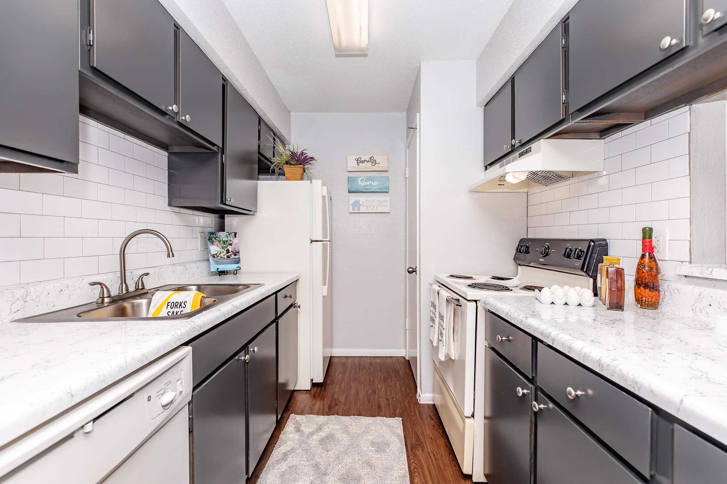 A modern kitchen featuring dark cabinetry, white tile backsplash, and a light-colored countertop. It includes a stainless steel sink, a white refrigerator, an oven, and various kitchen items like a bottle, eggs, and a few decorative pieces on the counter. The space is well-lit and organized.