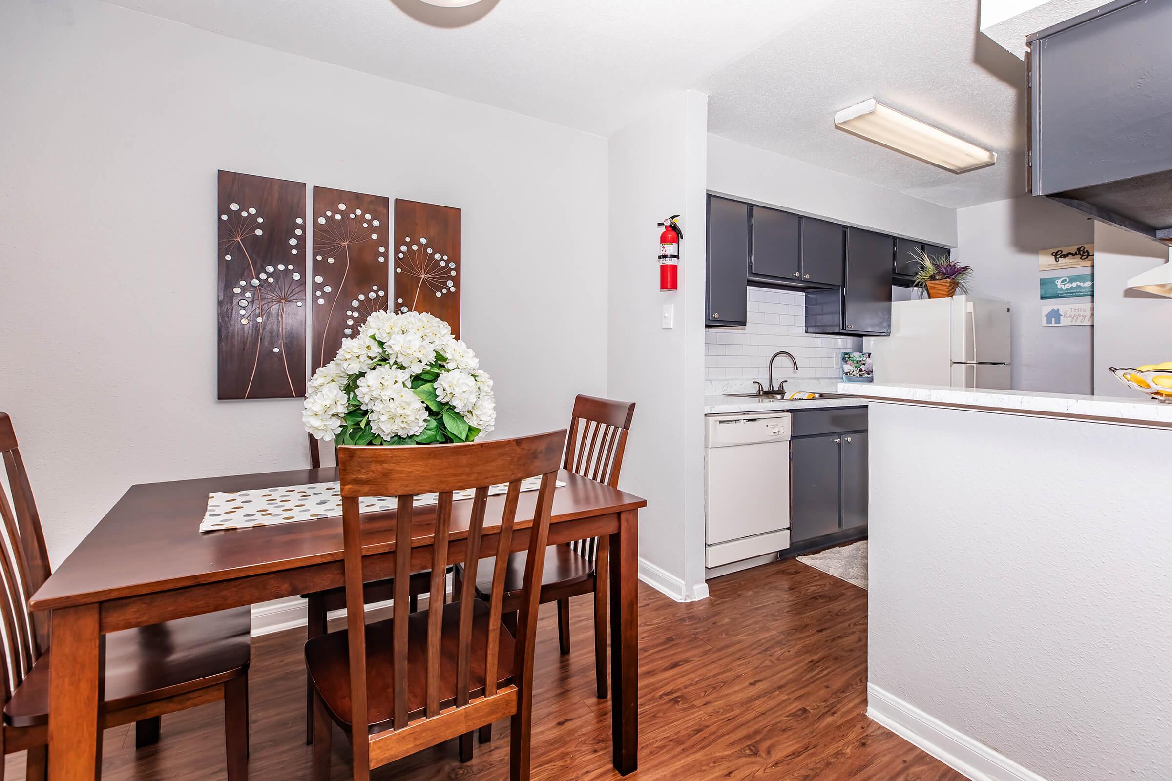 A bright dining area with a wooden table and chairs, featuring a floral centerpiece. In the background, a modern kitchen is visible with dark cabinets and a white countertop. The space is well-lit, with light-colored walls and a decorative wall art of white flowers.