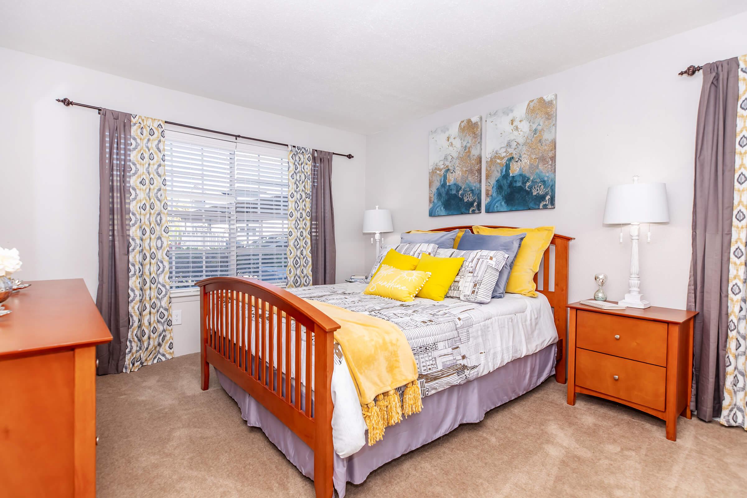 A cozy bedroom featuring a wooden bed with yellow and gray bedding, two decorative pillows, and a yellow throw. There's a matching wooden dresser and nightstand with lamps on each side. The windows are adorned with patterned curtains, and abstract artwork hangs above the bed, adding a touch of color to the serene atmosphere.