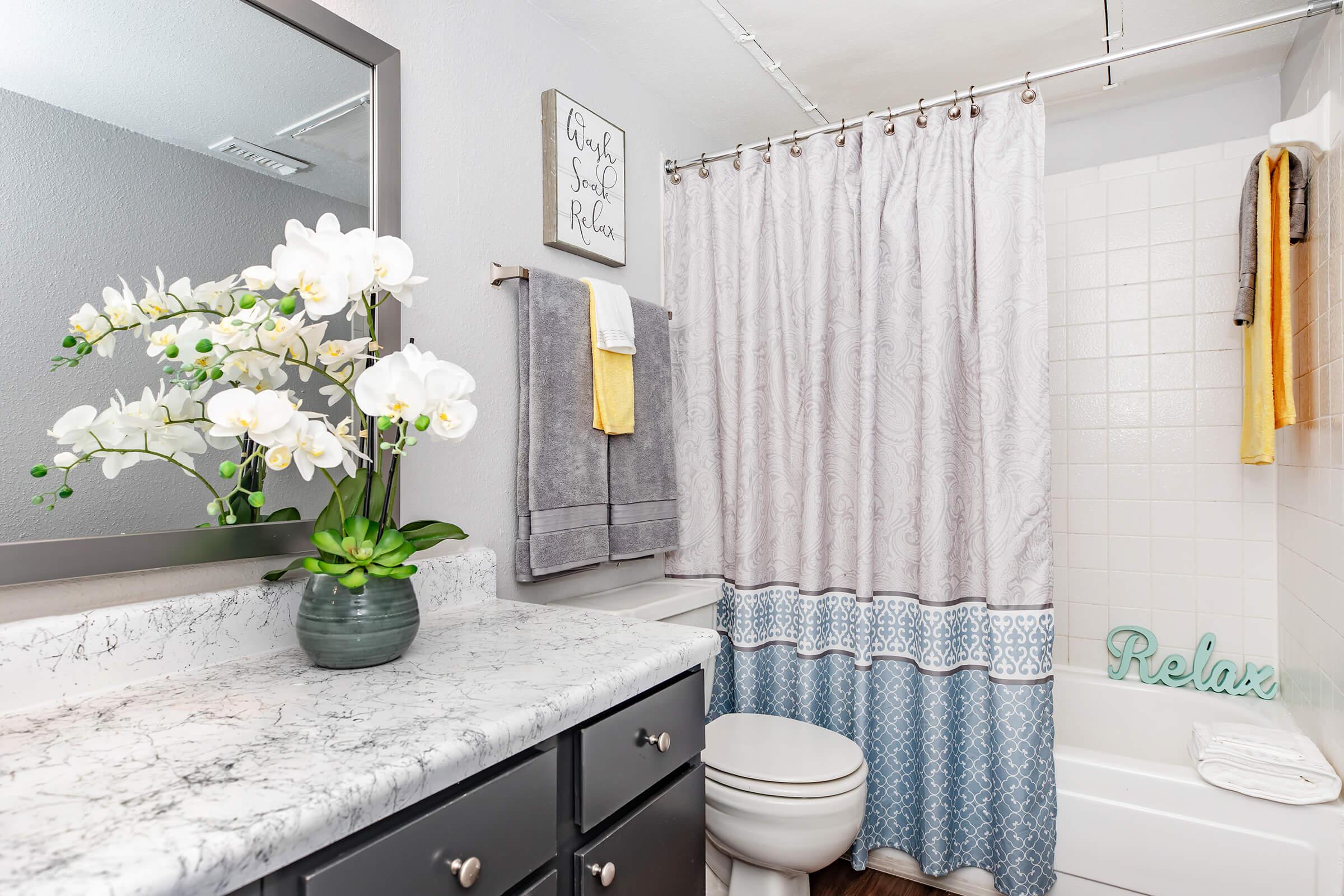 A clean bathroom featuring a marble countertop with a vase of white orchids, a mirror above the sink, a shower curtain with a blue and white pattern, towels hanging on a rack, and a relaxing decor piece that says "Relax" near the bathtub.