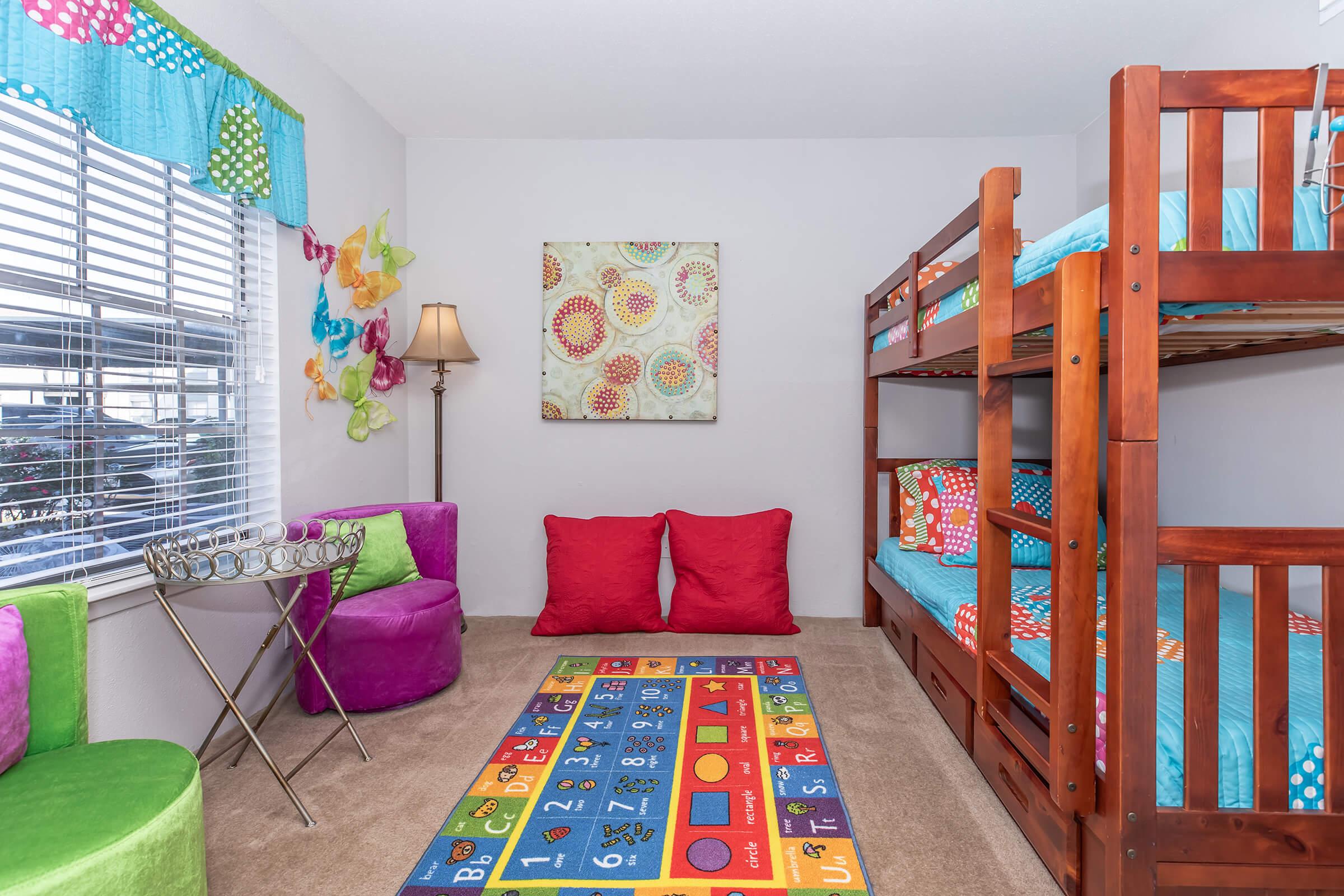 A colorful children's bedroom featuring a wooden bunk bed with blue bedding, a playful area rug with numbers, bright red floor cushions, a small decorative side table, and colorful wall decorations. Natural light streams in from a window with patterned curtains.