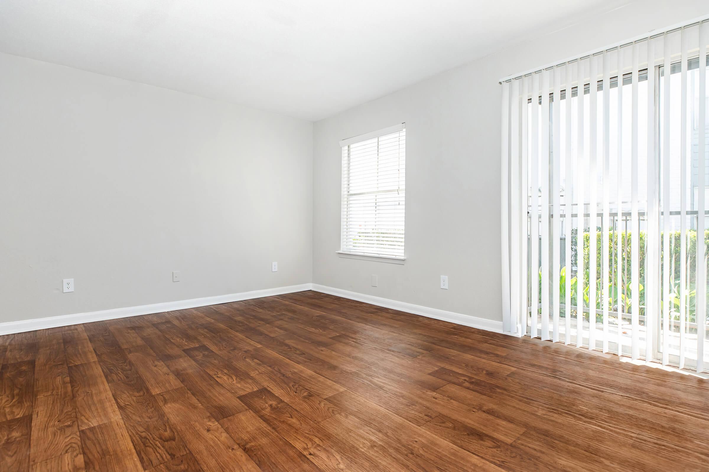 A bright, empty room with light gray walls and wooden flooring. A large window with white vertical blinds allows natural light to fill the space. The window is positioned next to a sliding glass door that opens to an outdoor area. The overall atmosphere is clean and welcoming, ideal for furnishing.