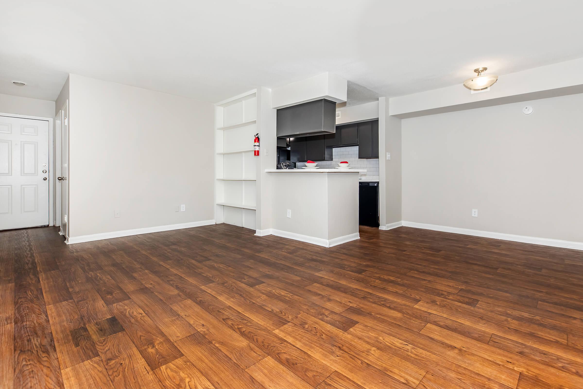 Spacious interior of a modern living area with hardwood flooring, featuring a shelf unit and an open kitchen in the background. The kitchen includes dark cabinets and white countertops. A doorway leads outside, with a fire extinguisher visible on the wall. Bright and neutral-colored walls enhance the airy feel.