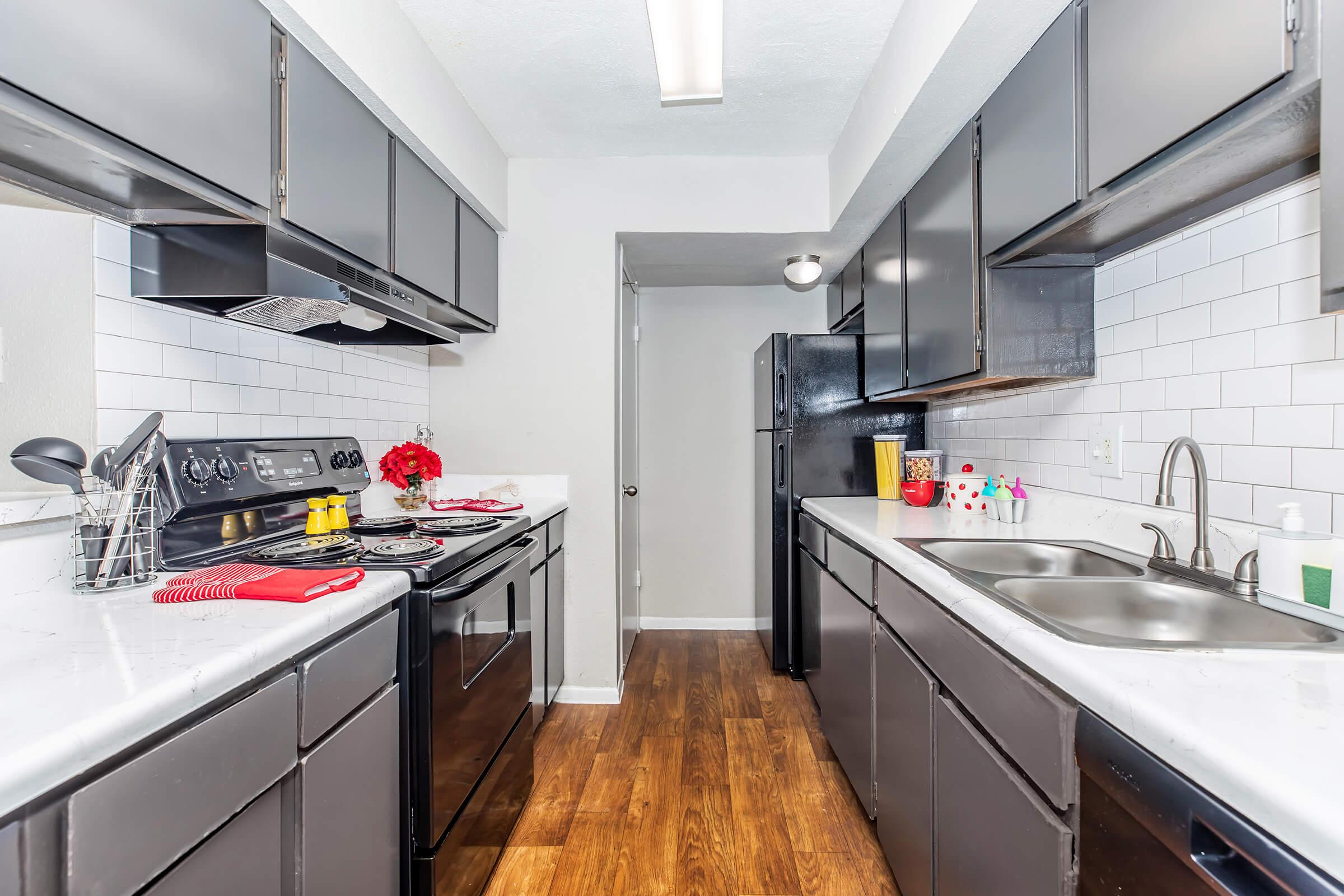 A modern kitchen featuring dark cabinetry, white subway tile backsplash, and stainless steel appliances. The countertops are adorned with decorative items, and the flooring is a warm wood finish. The layout includes an open space between two countertops, leading to a doorway at the far end.