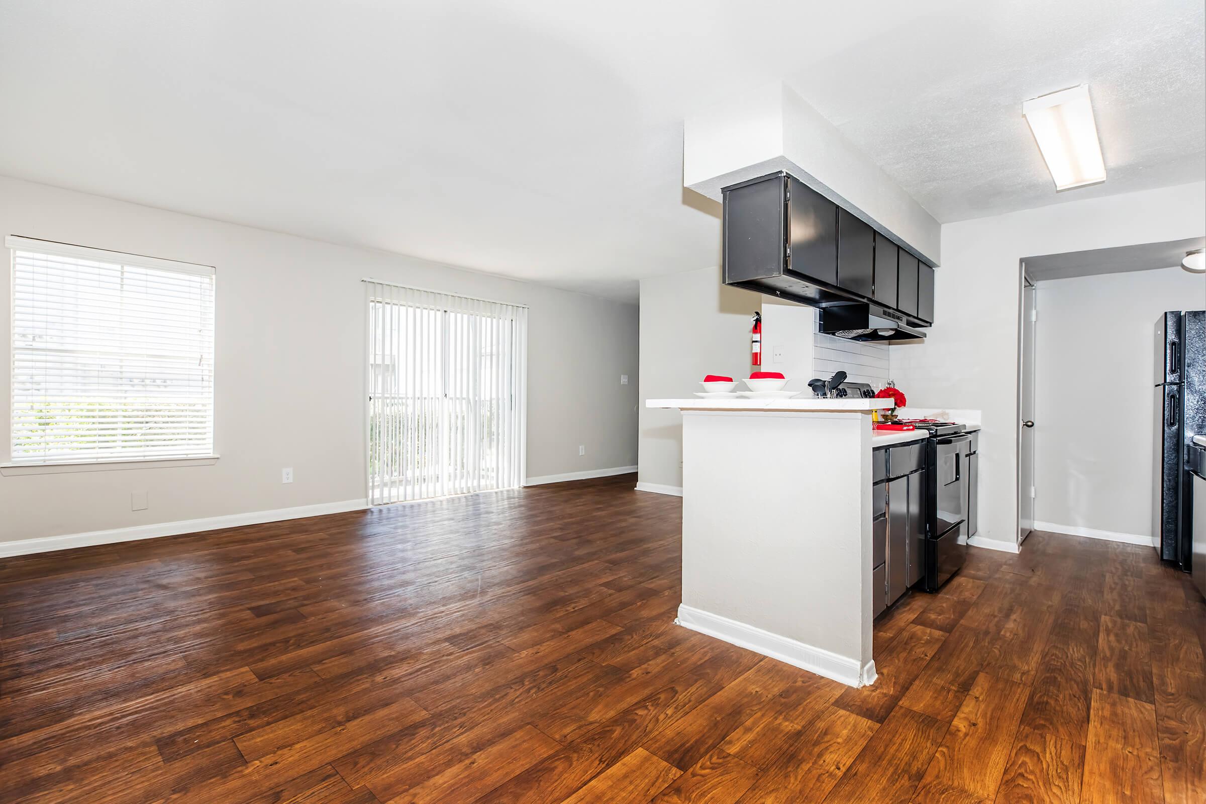 A spacious, modern kitchen and living area with hardwood flooring. The kitchen features dark cabinets, a gas stove, and a countertop with red cookware. Natural light streams in through large windows with blinds in the living area, enhancing the bright and inviting atmosphere.