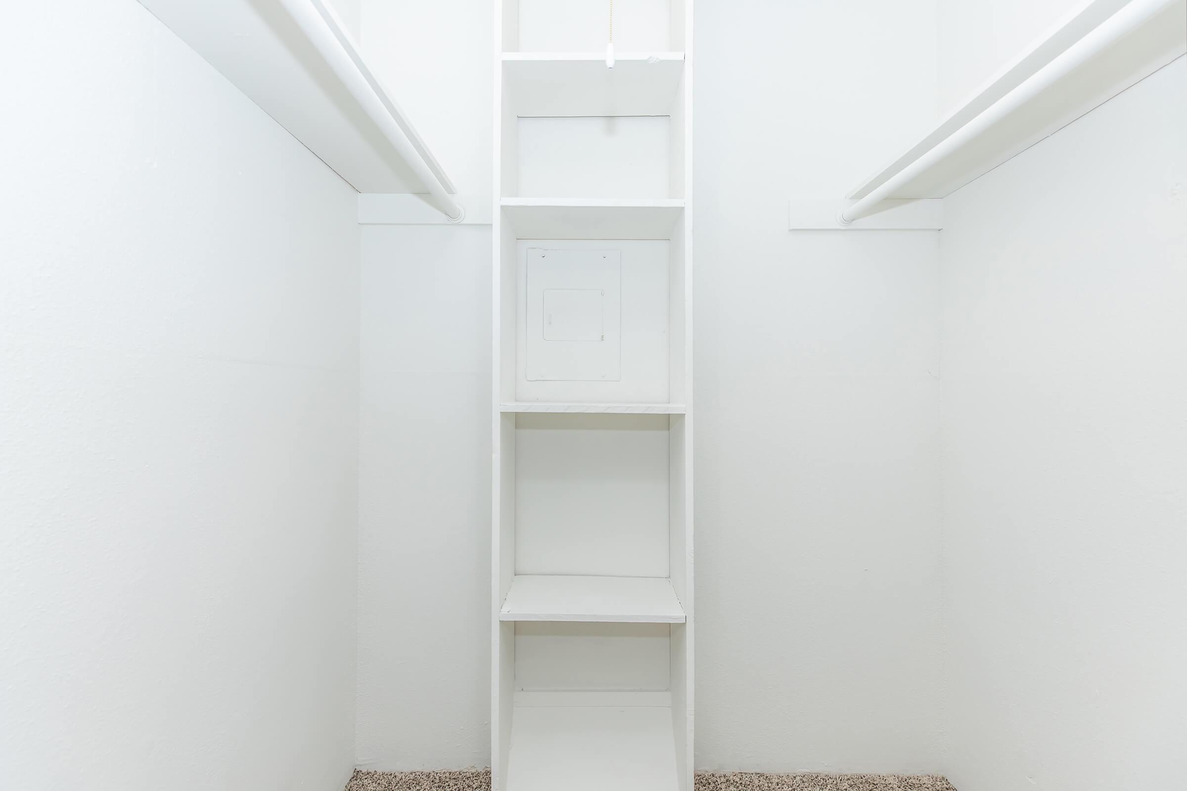 A bright, empty closet featuring white walls and shelving. The shelving is arranged vertically in the center, with a small access panel visible. The floor is covered in a light textured carpet, creating a clean and minimalist appearance.