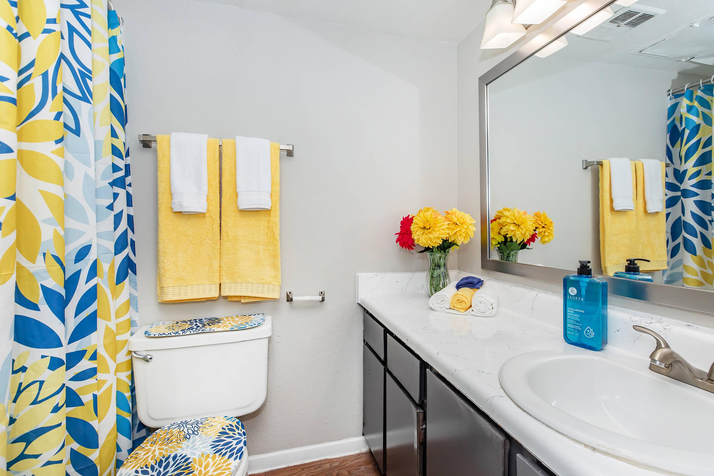 A bright and cheerful bathroom featuring a decorative yellow and blue shower curtain, white towels hanging on a towel rack, and a vanity with a white sink. There are colorful flowers in a vase on the countertop, and toiletries neatly arranged. The wall is painted a light gray.
